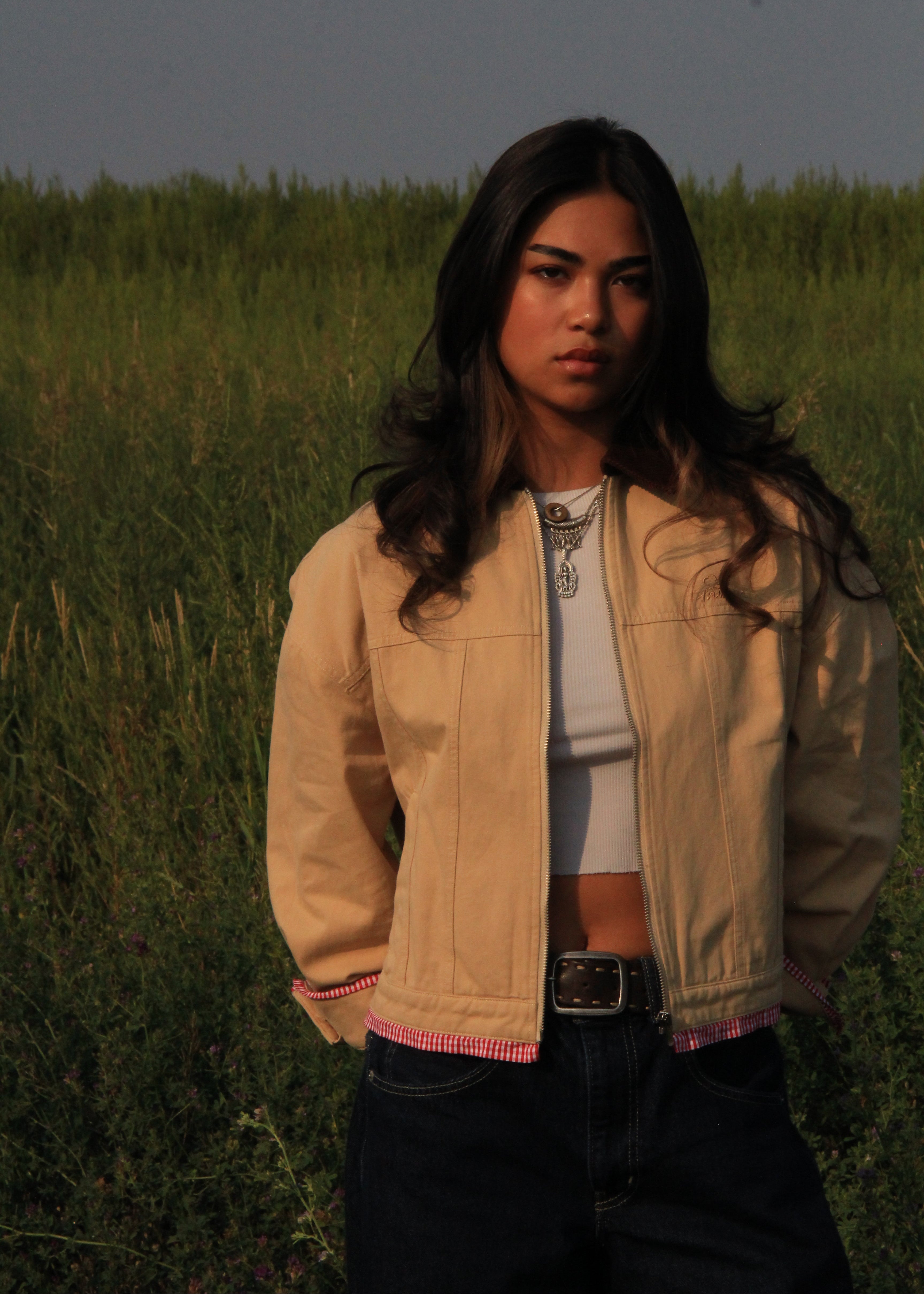 A woman standing in a grassy field wearing a beige cropped jacket over a white crop top and dark jeans.