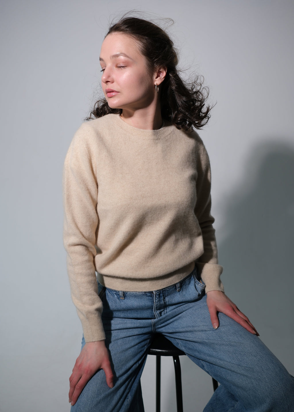 A woman wearing a beige crewneck knit sweater and blue jeans sits on a black stool against a plain gray studio background.