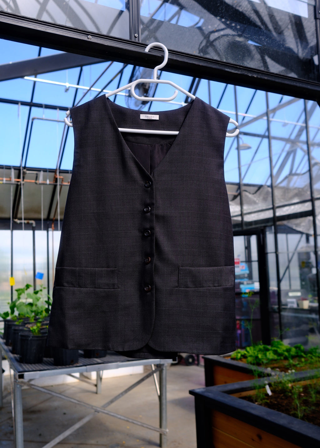 A black sleeveless vest with a buttoned front and notched lapels, hanging on a white hanger in an outdoor patio setting with glass windows and plants visible.