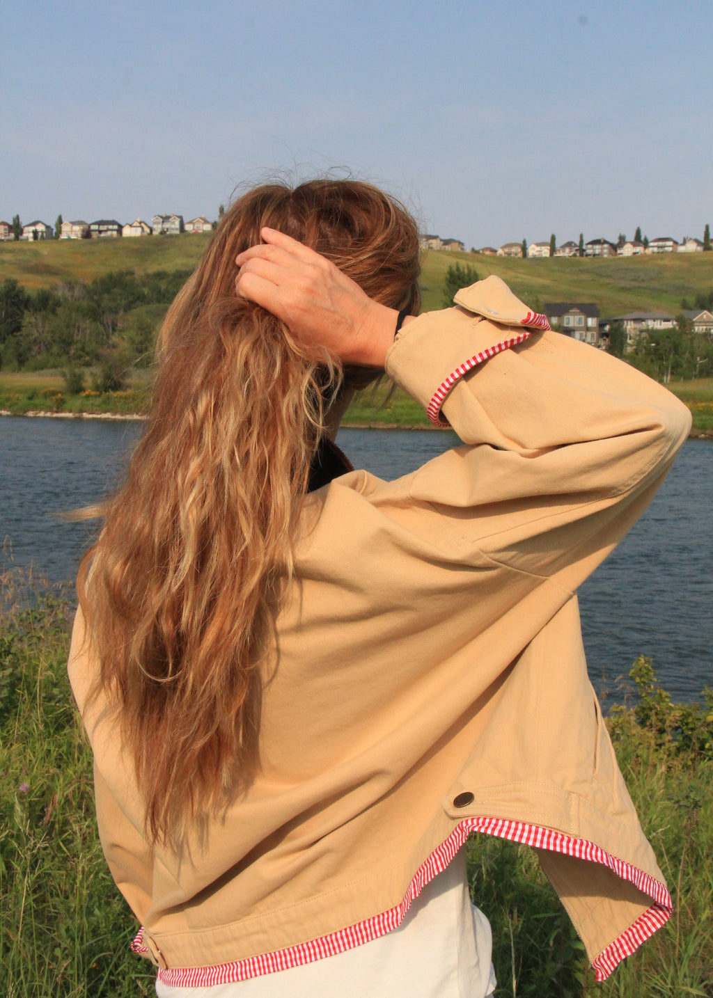 A woman with long wavy light brown hair stands outdoors by a lake, seen from behind, wearing a beige oversized jacket with red and white gingham trim over a white top.