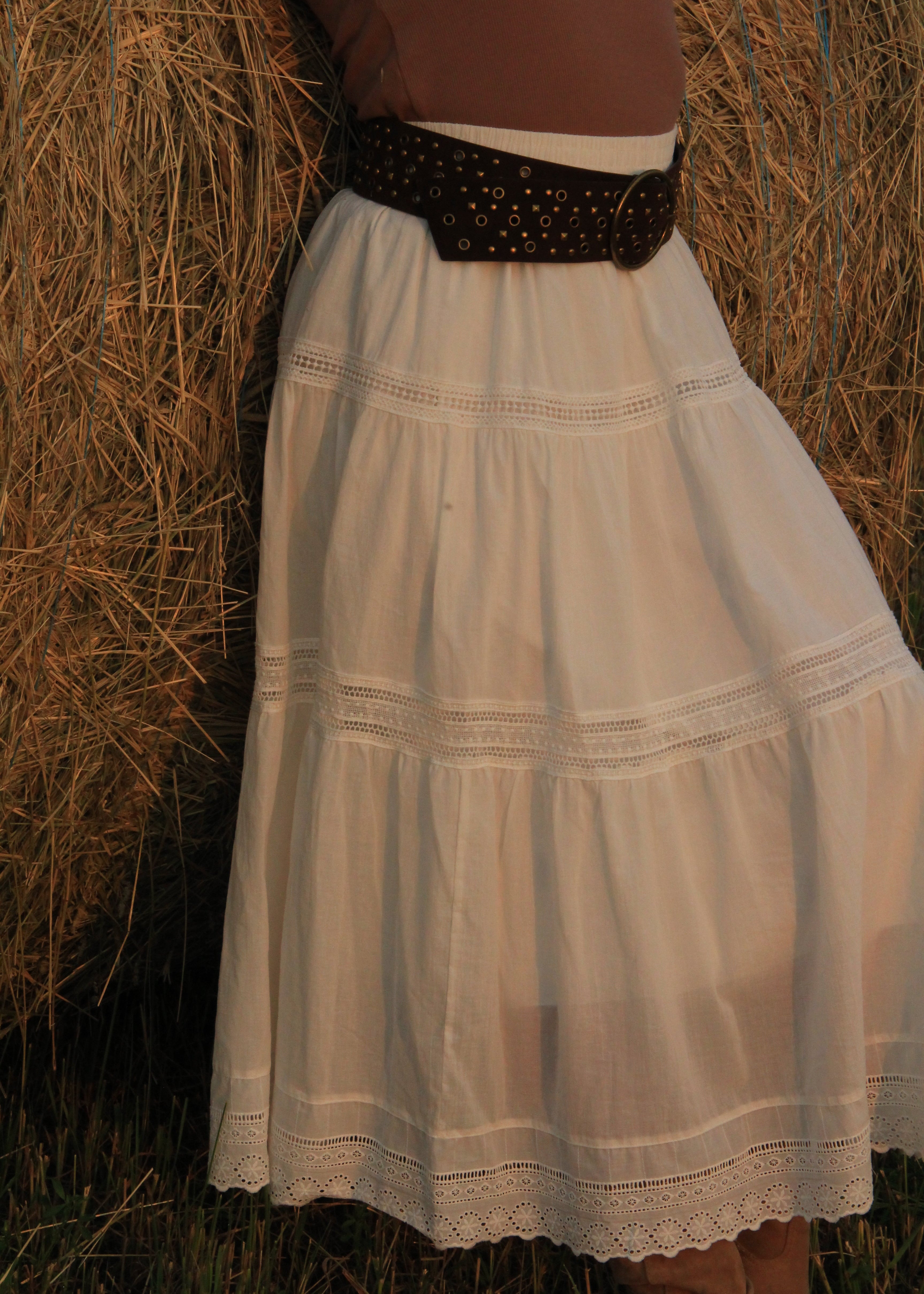 A white tiered cotton skirt with delicate lace trim and a wide black studded belt, shown on a model standing in front of a hay bale.