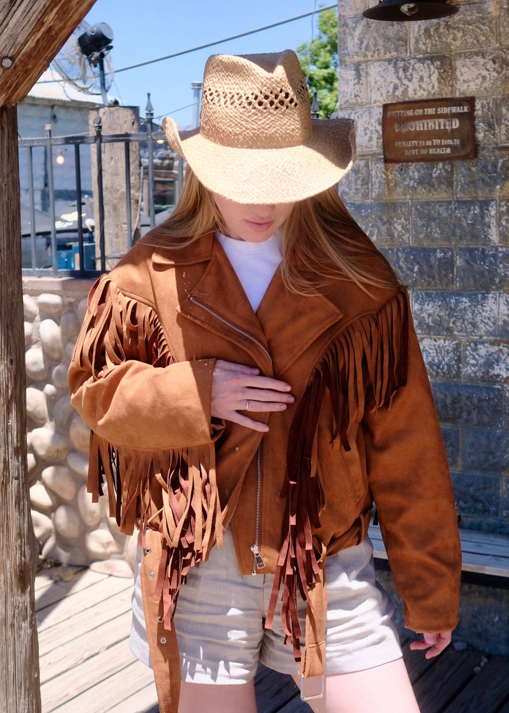 Person wearing a brown fringed jacket and straw hat in front of a rustic building.