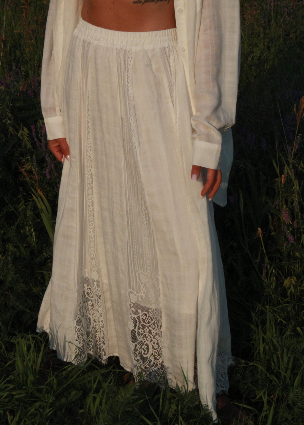 A person outdoors in a field wearing a long white linen maxi skirt with lace trim at the hem, paired with a matching white shirt.