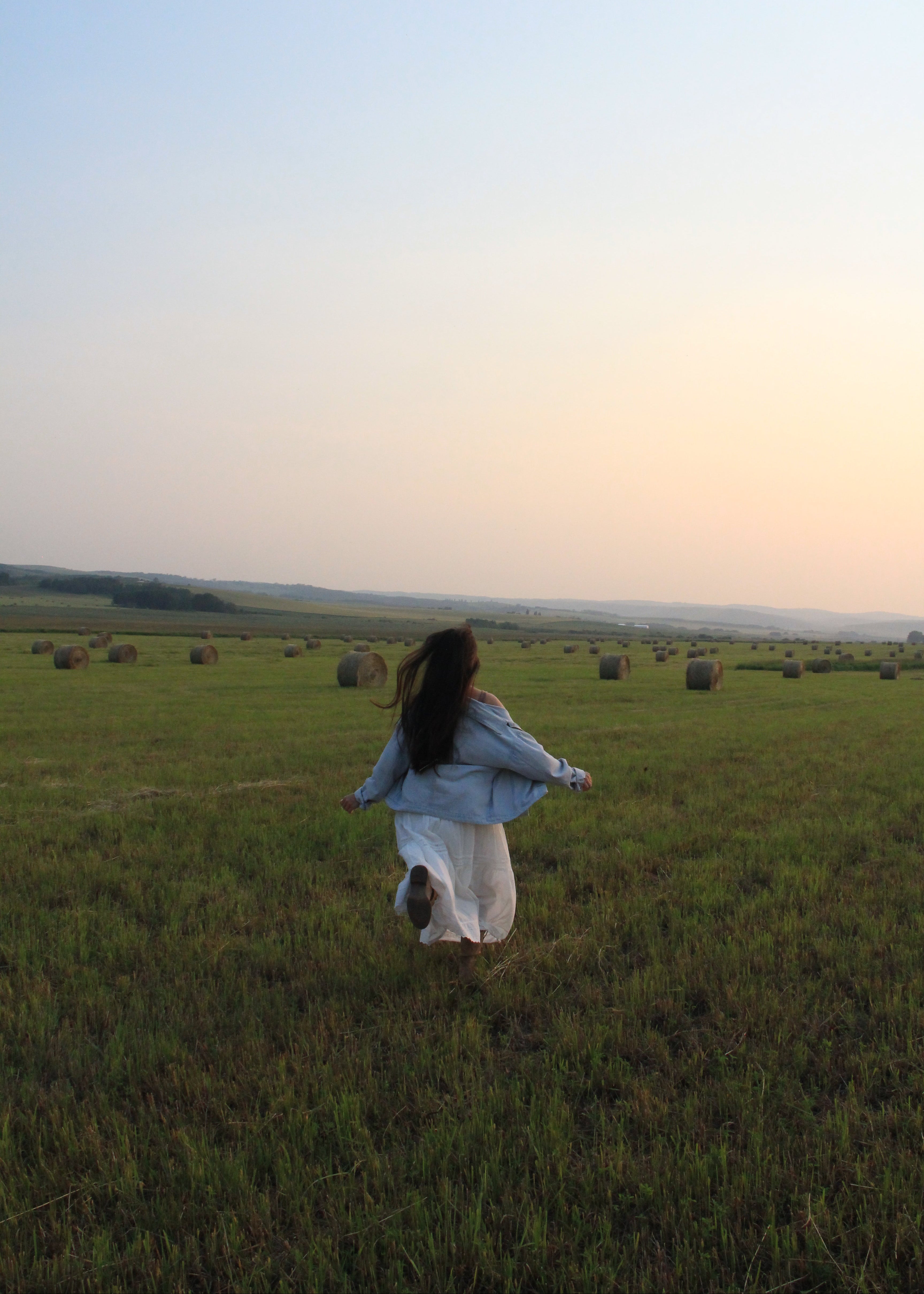A woman in a light blue denim jacket and white skirt runs joyfully across a grassy field with hay bales under a soft sunset sky.