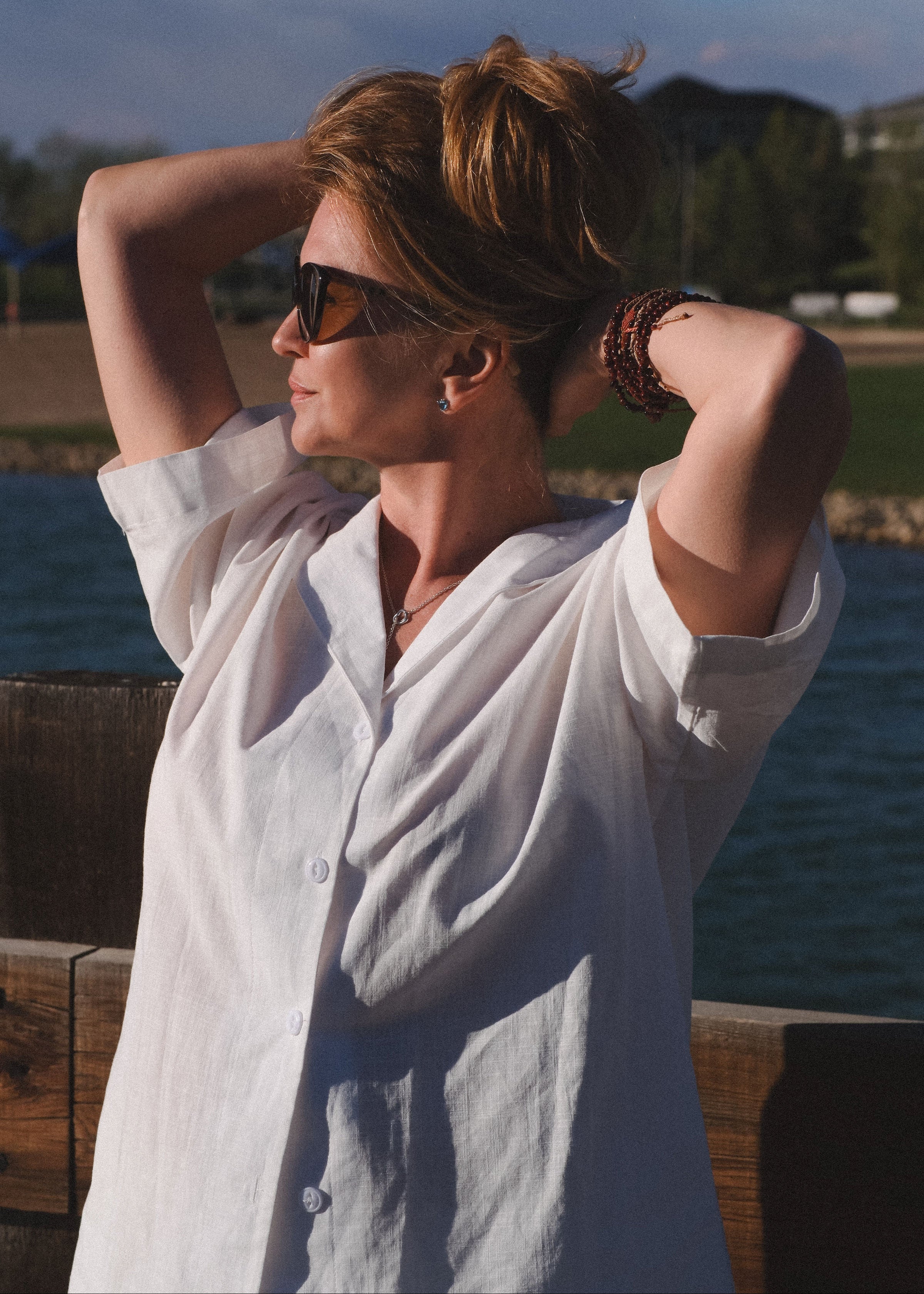 Woman in a white shirt with sunglasses, standing by a body of water.