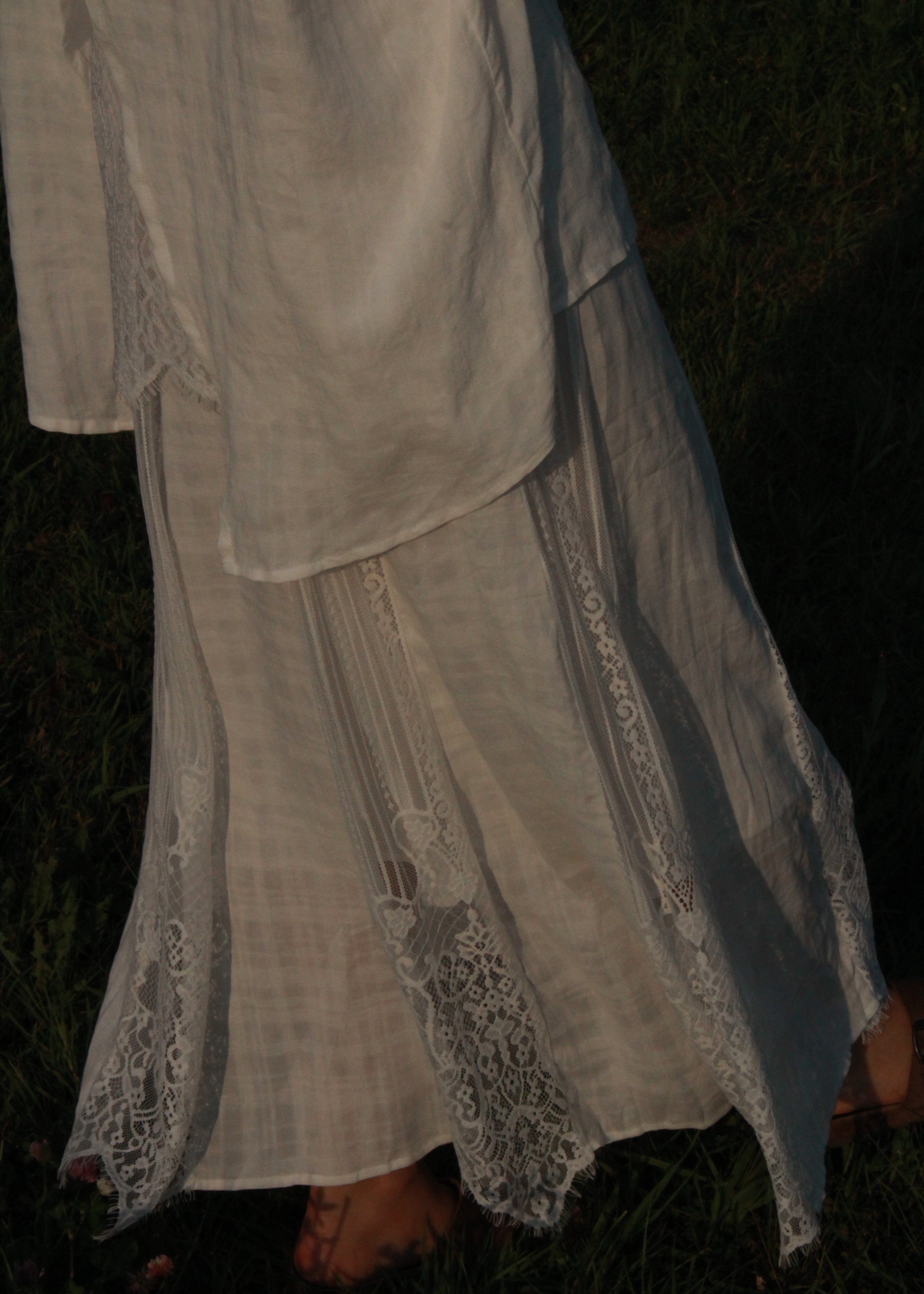 A white or off-white long-sleeve dress or robe with delicate lace trim along the cuffs and front, shown draped outdoors on dark grass.