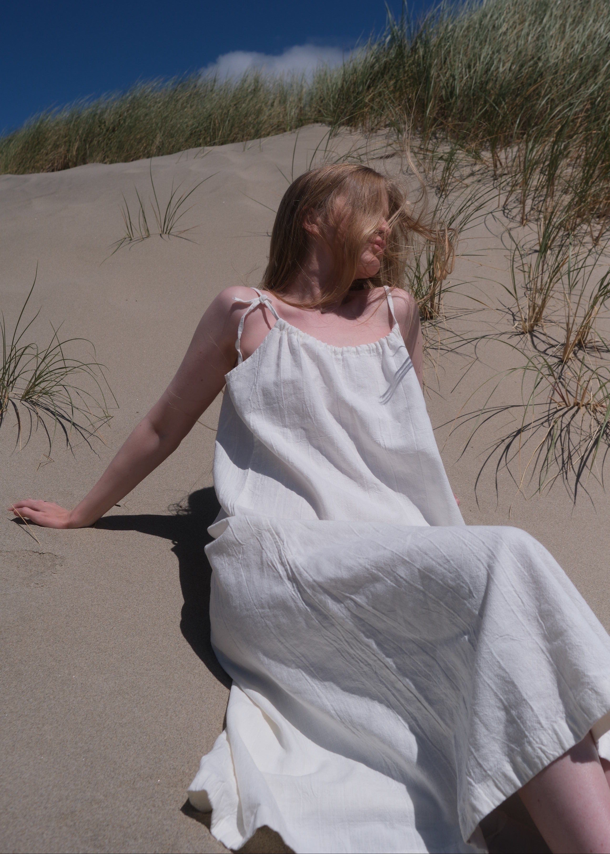 Person in a white linen  dress standing on a sandy beach with blue sky.