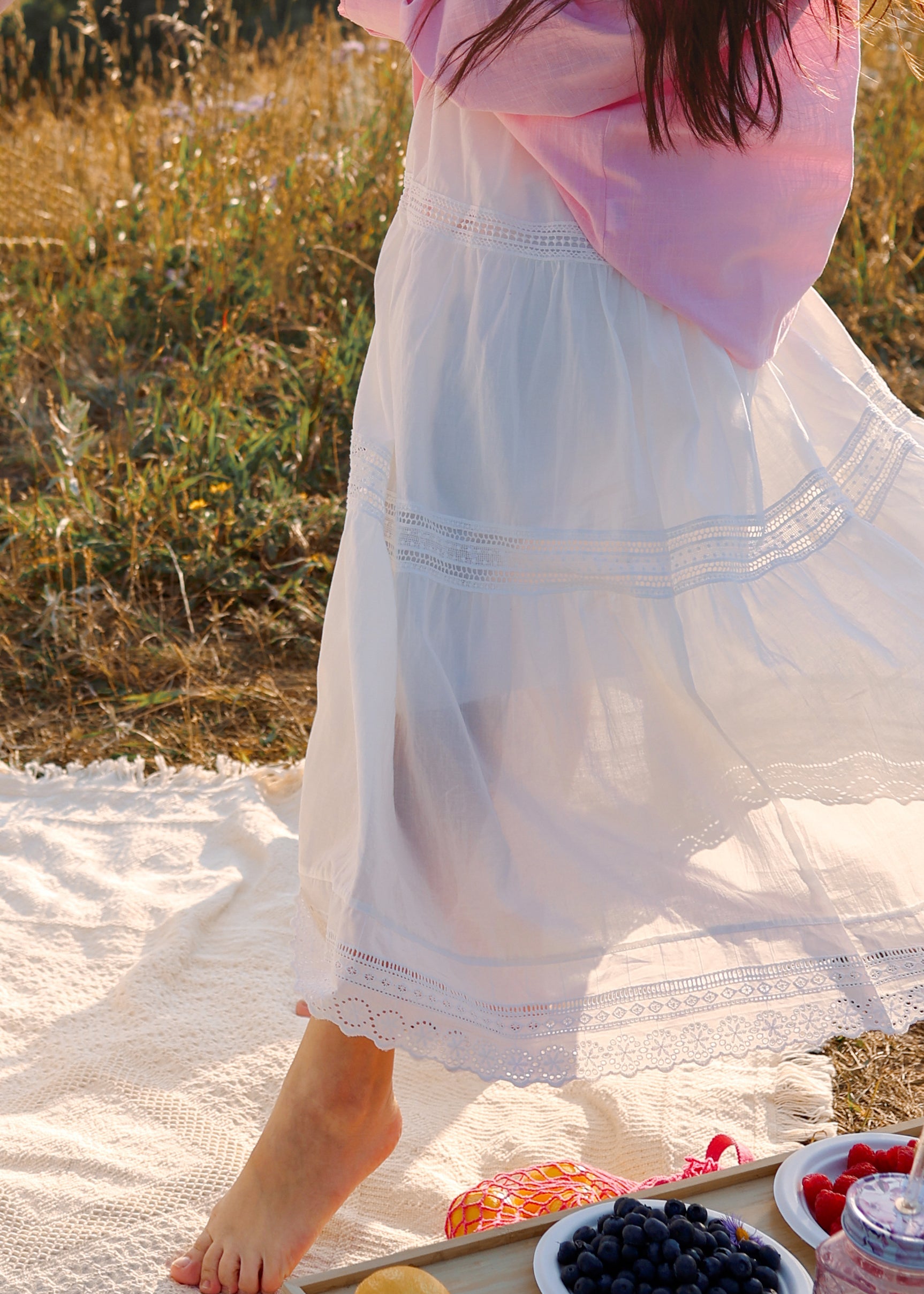 A person outdoors on a white picnic blanket, wearing a white tiered dress with a light pink top. A wooden tray holds fruit and a bowl of blueberries in a sunlit field.