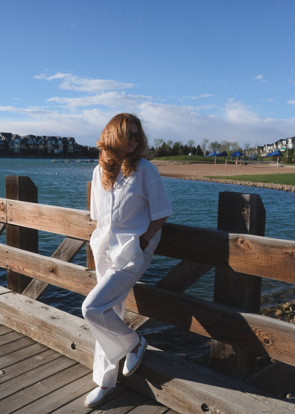 Person in white outfit sitting on a wooden bench by a waterfront with a blue sky.