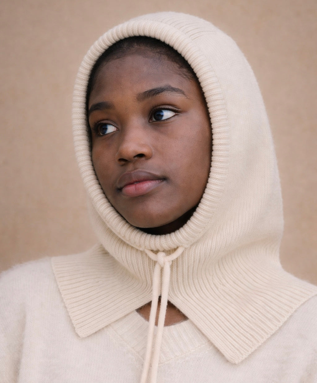 Person wearing a beige knitted hood with a high collar against a beige background