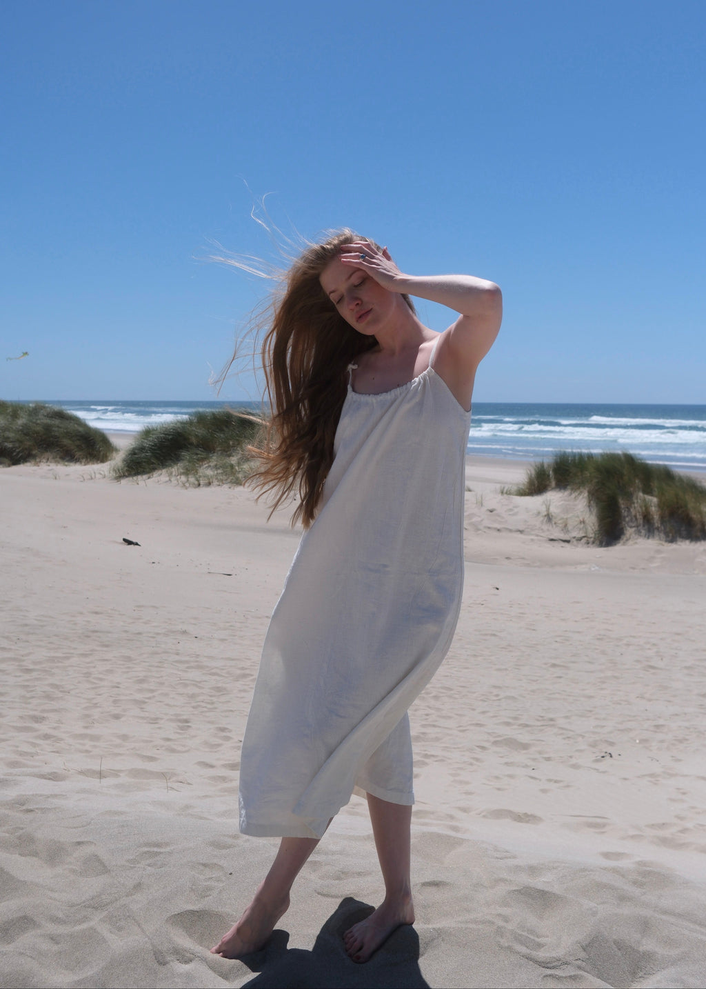 Woman in a white dress standing on a sandy beach with a clear blue sky.