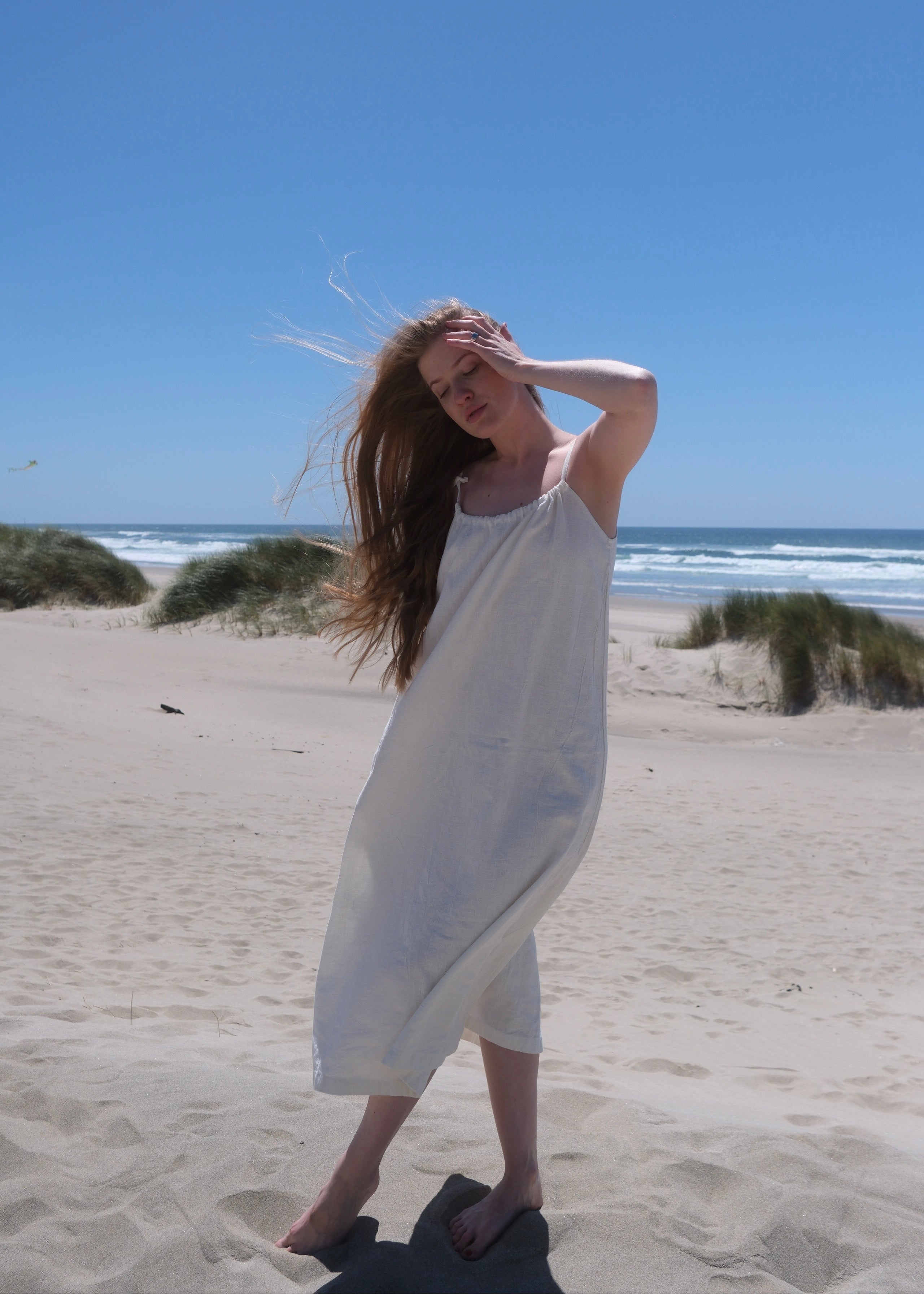 Woman in a white dress standing on a sandy beach with a clear blue sky.