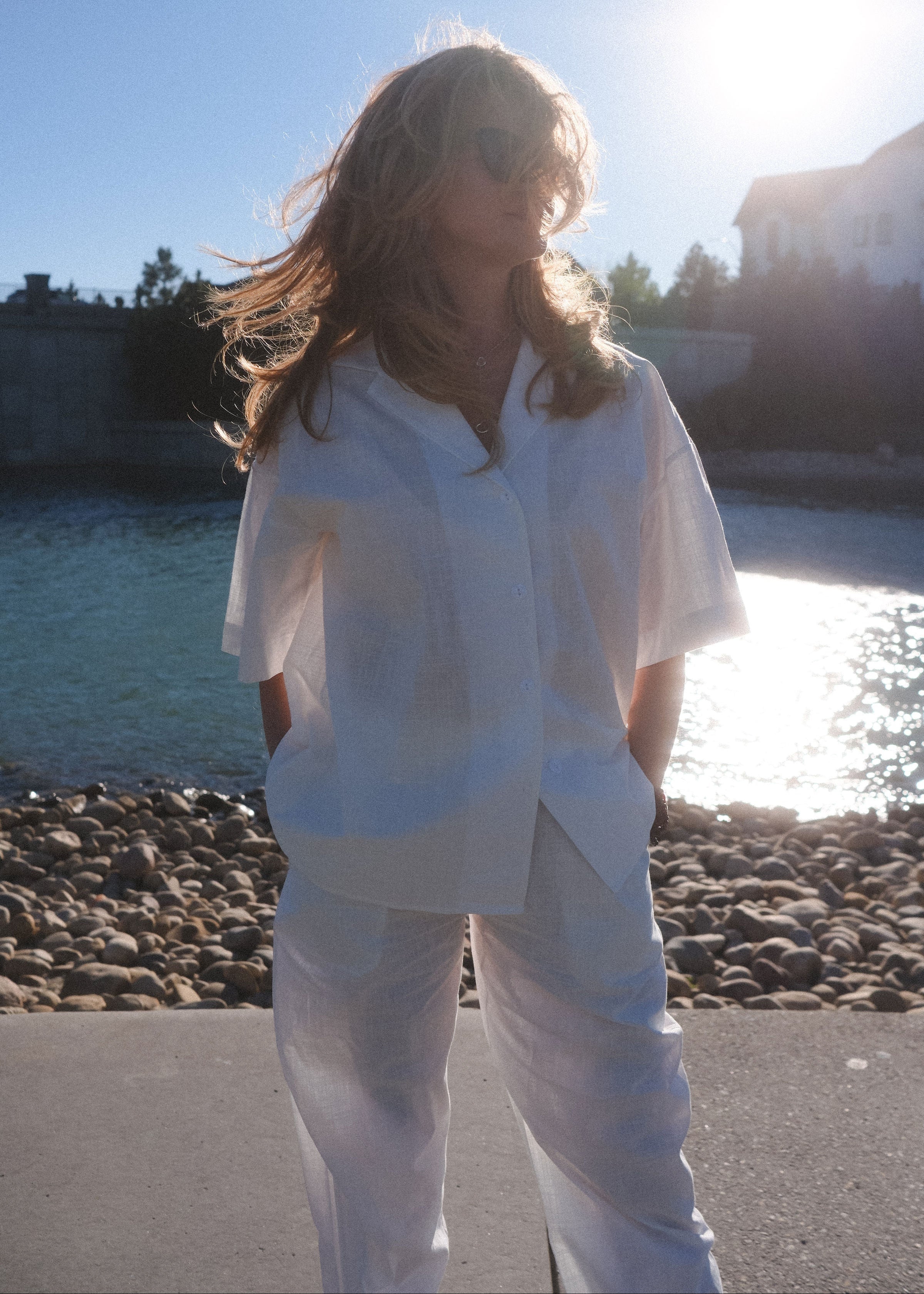 Person wearing a white outfit standing by a body of water with trees and buildings in the background.
