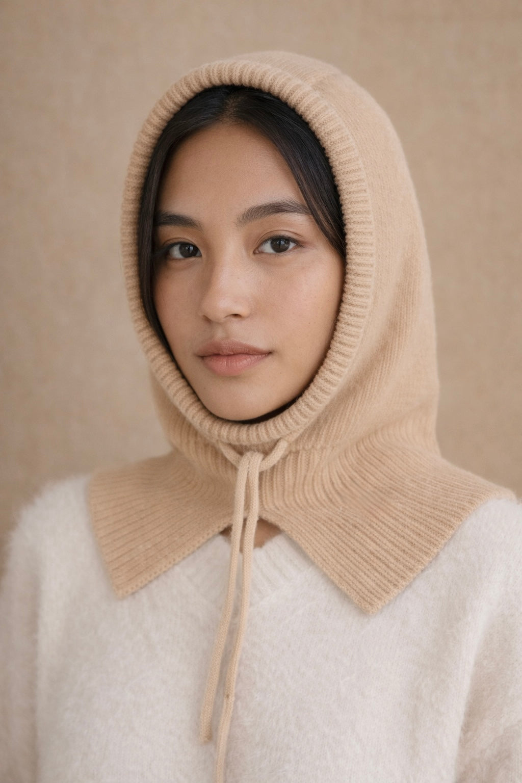Person wearing a beige knitted hood over a white garment against a beige background