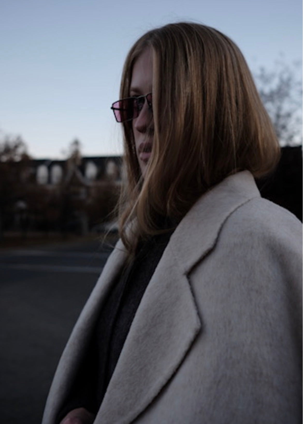 A woman with shoulder-length light brown hair and dark sunglasses, wearing a light beige wool coat over a dark top, photographed outdoors in low light.