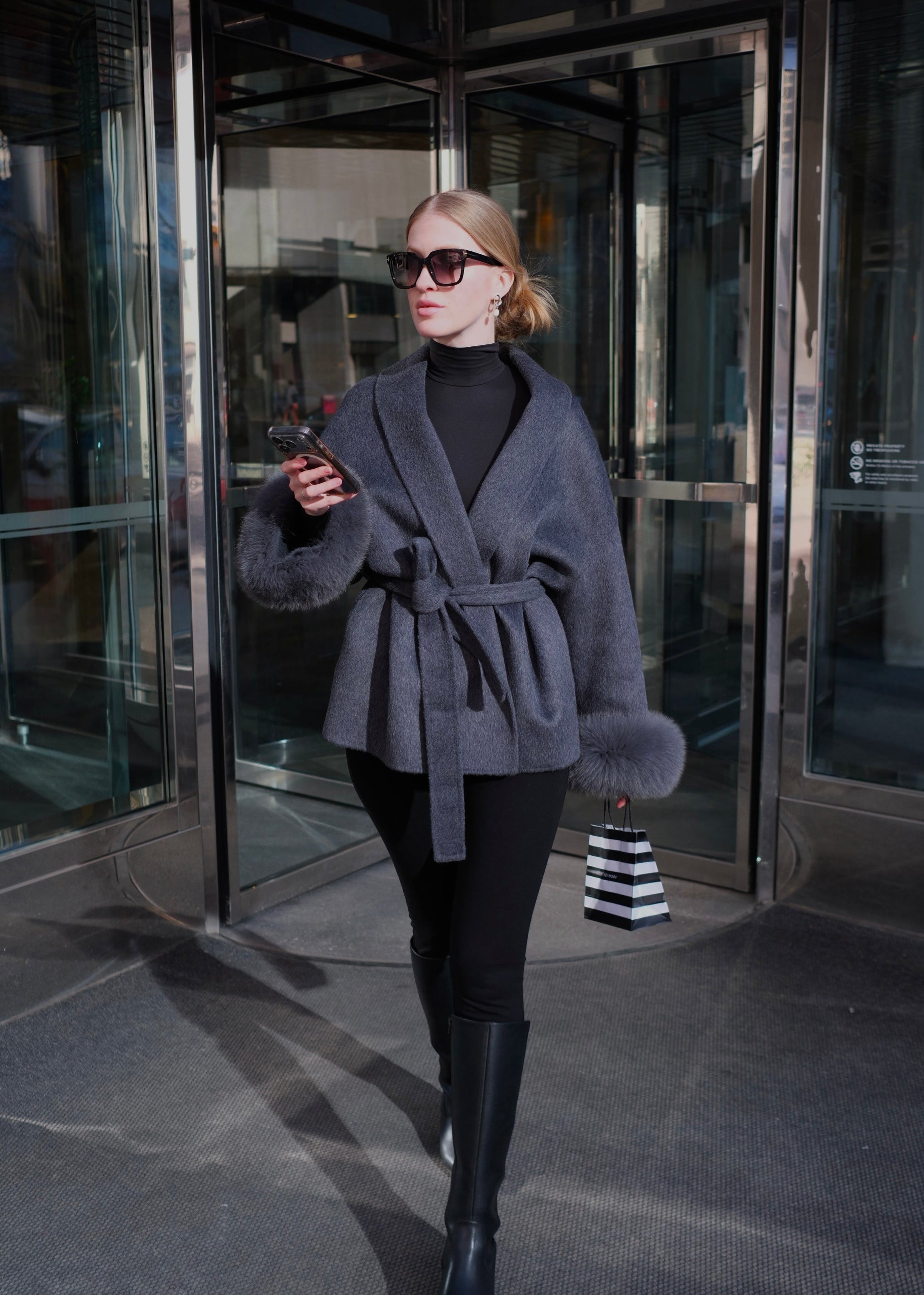 Woman in a gray coat and black pants walking out of a building.