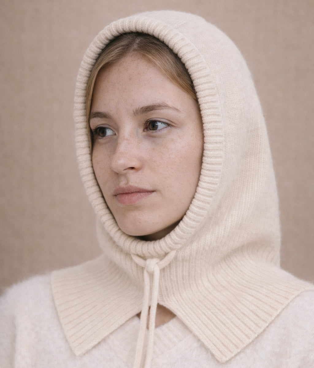 Person wearing a cream-colored knitted hood against a beige background