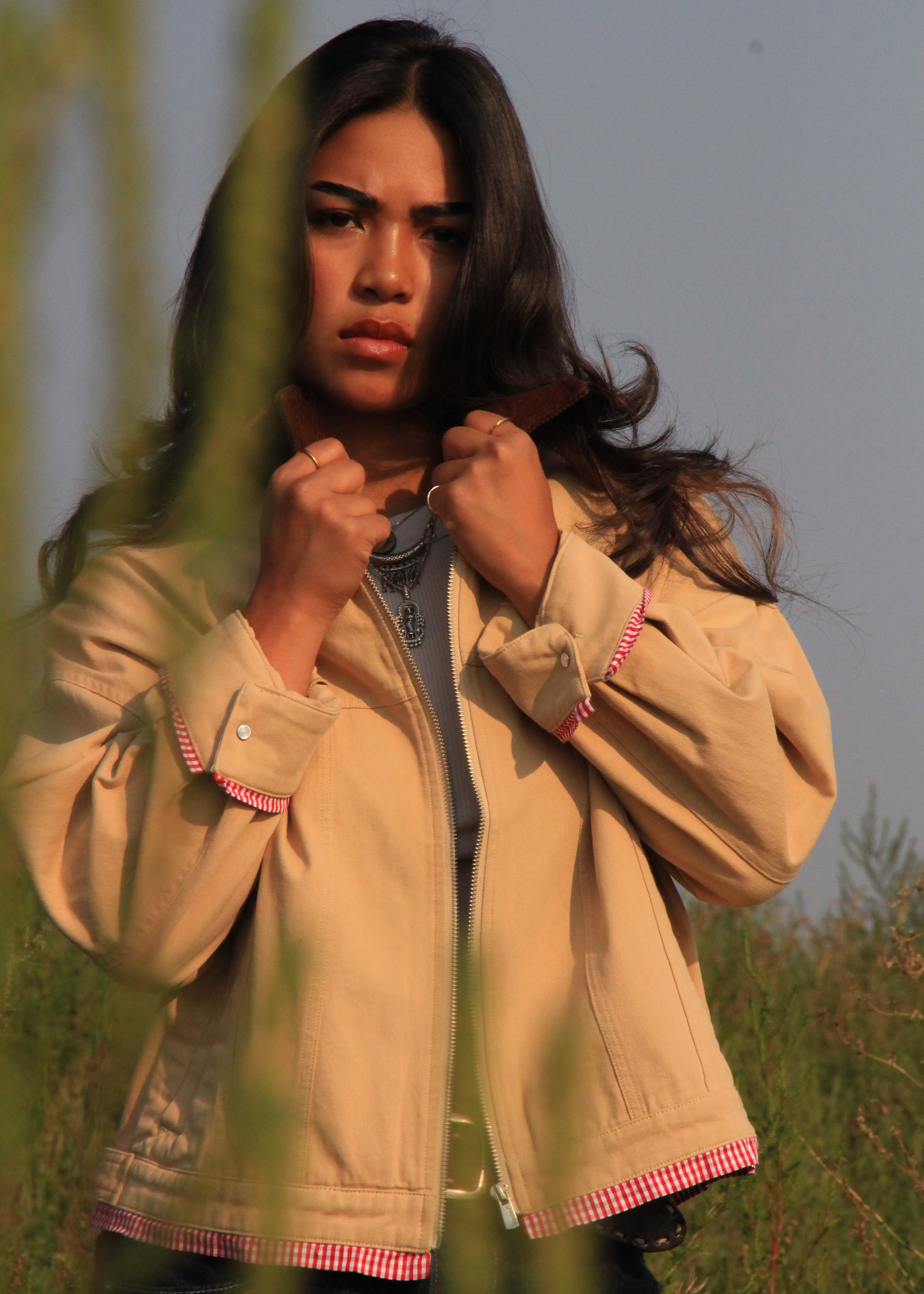 A woman wearing a beige jacket with pink and white striped trim, standing outdoors in a grassy field.