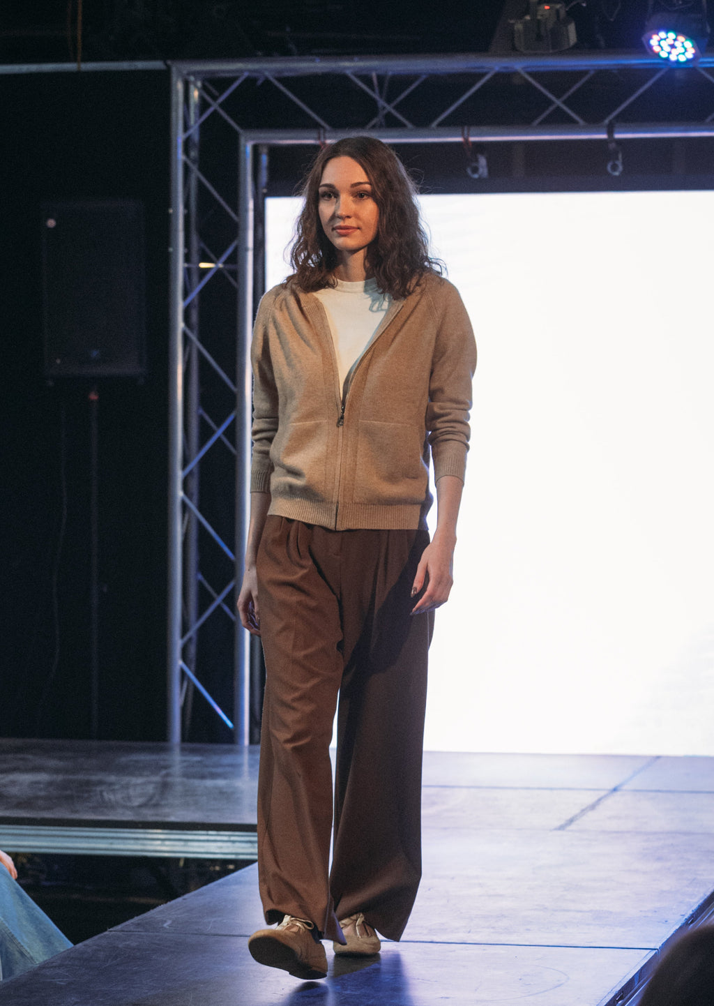 A model walks a fashion runway wearing a beige zip-up cardigan over a white top and brown wide-leg trousers, with an audience seated along the runway in a dark studio setting.