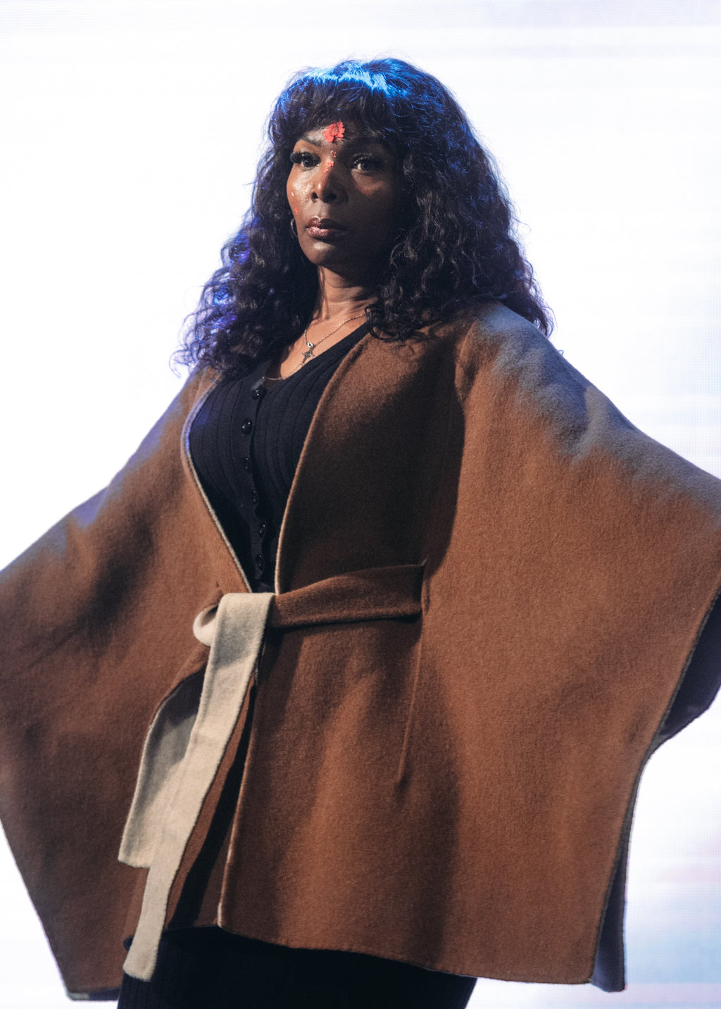 A model with long curly dark hair stands in a studio wearing a brown draped cape-style coat with a cream tie belt over a black outfit, posed against a bright white background with soft pink and blue light streaks.