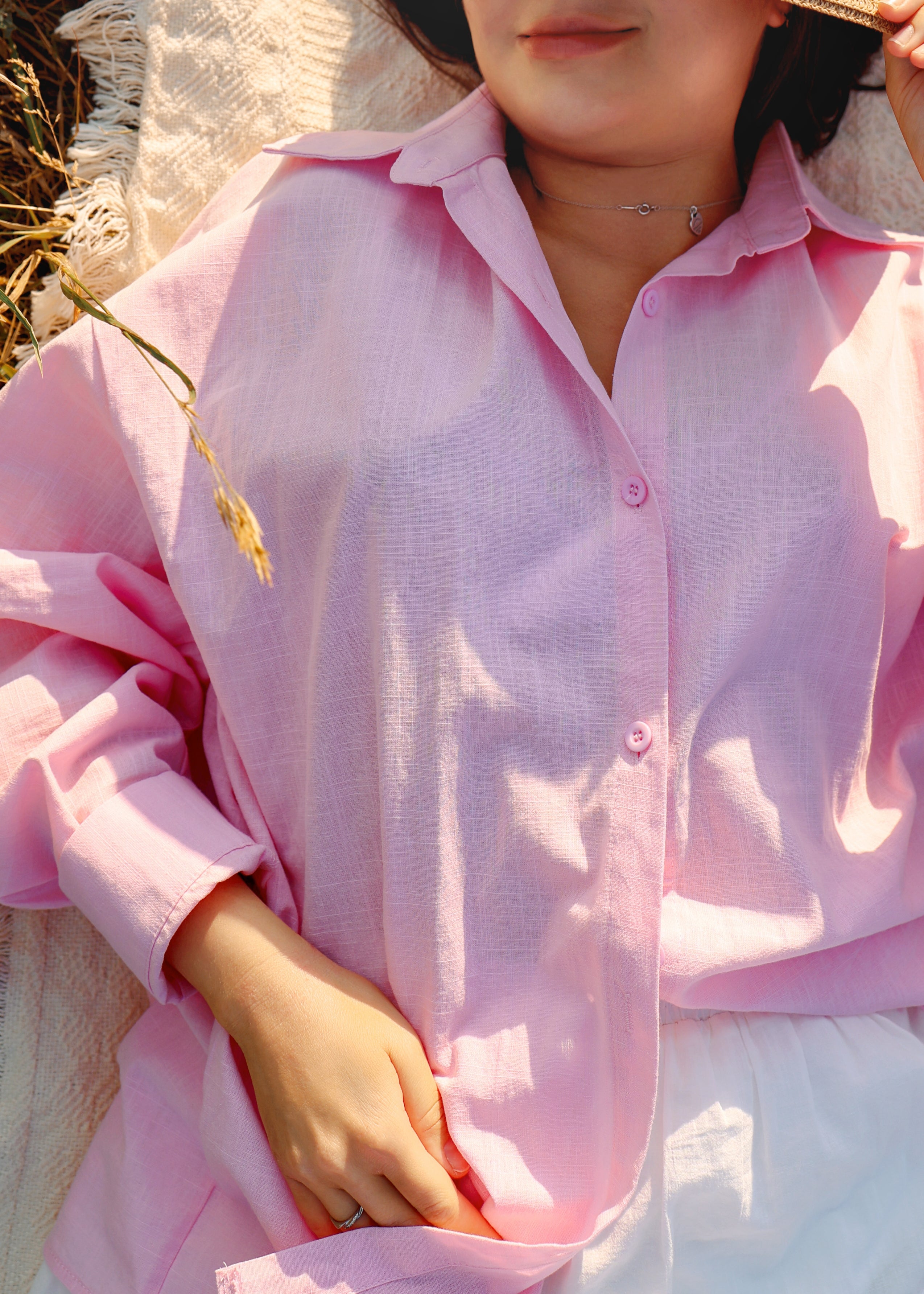 A close-up of a person wearing a light pink button-up shirt with a relaxed, oversized fit. The photo is taken in warm, sunny outdoor light, showing the fabric texture and soft drape.