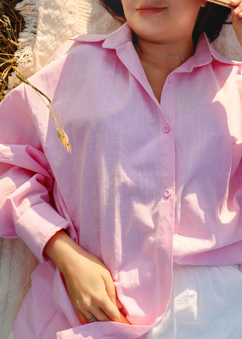 A close-up of a person wearing a light pink button-up shirt with a relaxed, oversized fit. The photo is taken in warm, sunny outdoor light, showing the fabric texture and soft drape.