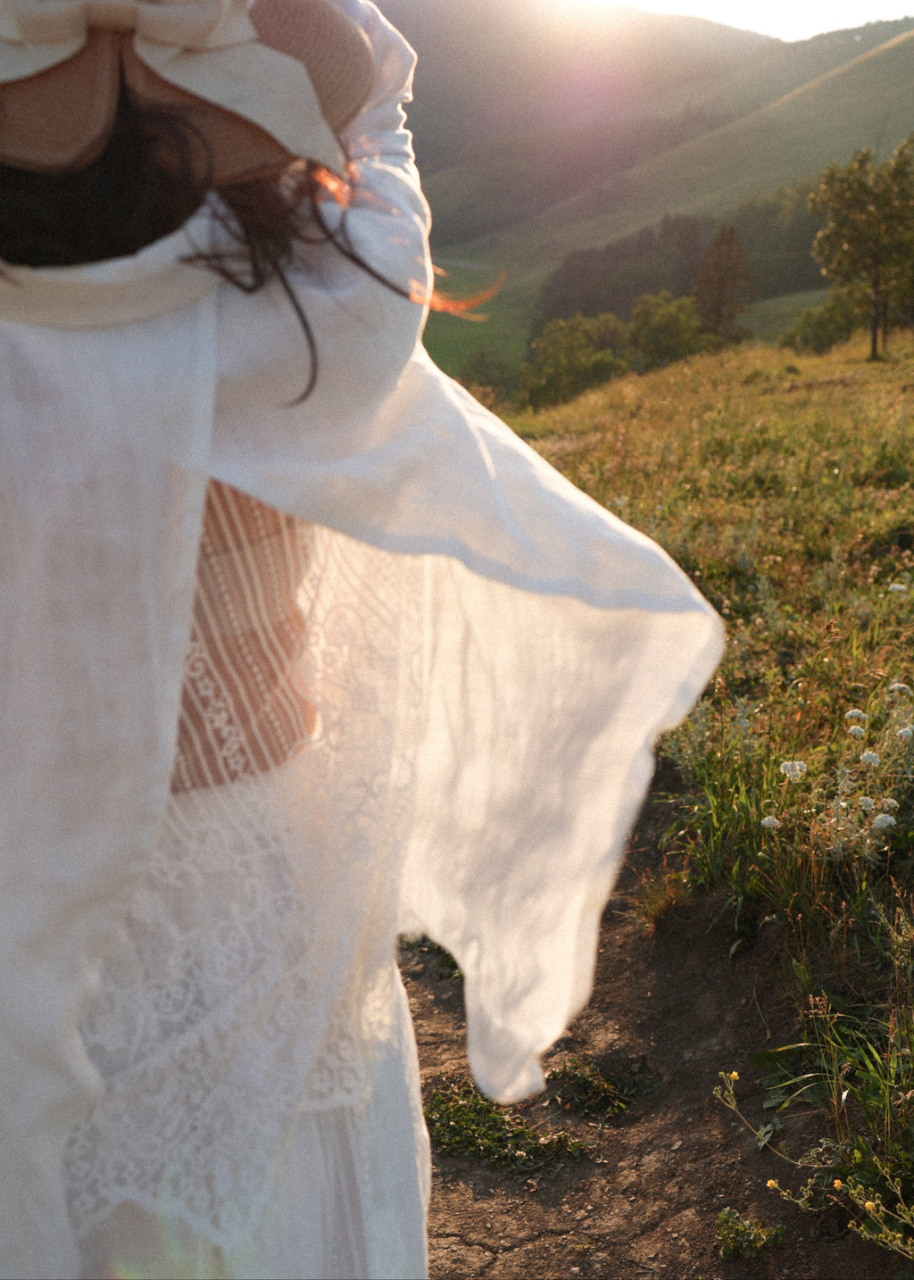 A woman outdoors in a sunlit grassy landscape, wearing a flowing white lace dress or robe with a large bow on the head. The warm sunlight creates a bright flare in the background with hills and trees.
