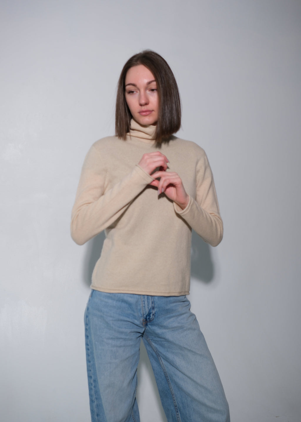A woman with shoulder-length brown hair wearing a beige ribbed turtleneck sweater and light blue wide-leg jeans, standing against a plain light gray wall.