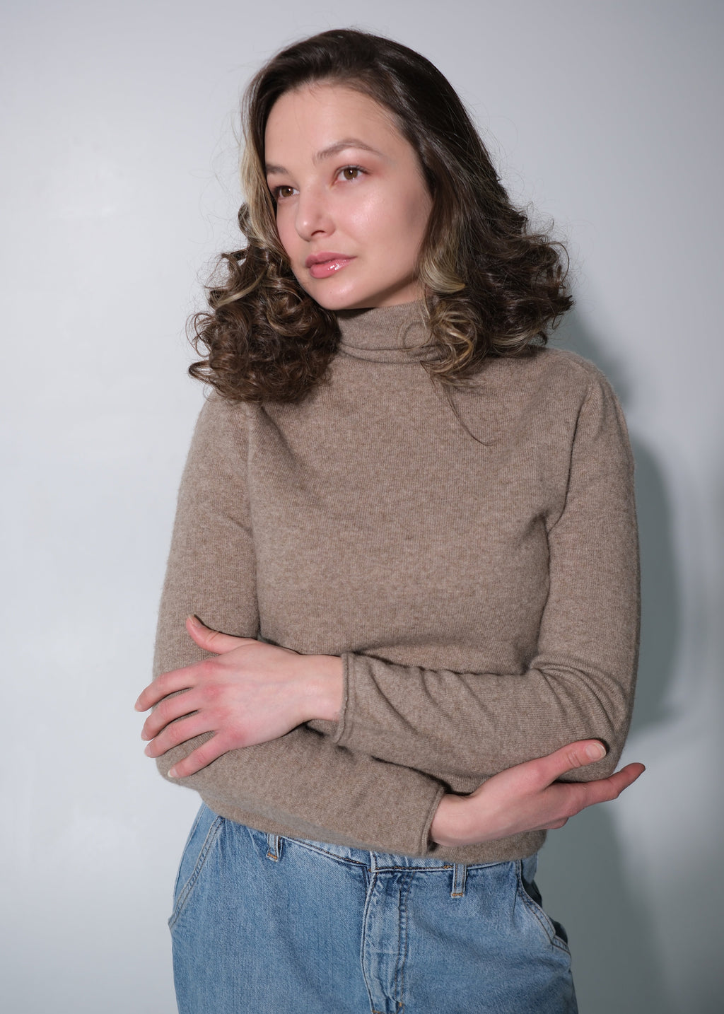 A woman with wavy brown hair wearing a beige turtleneck sweater and blue jeans, standing against a plain light gray background.