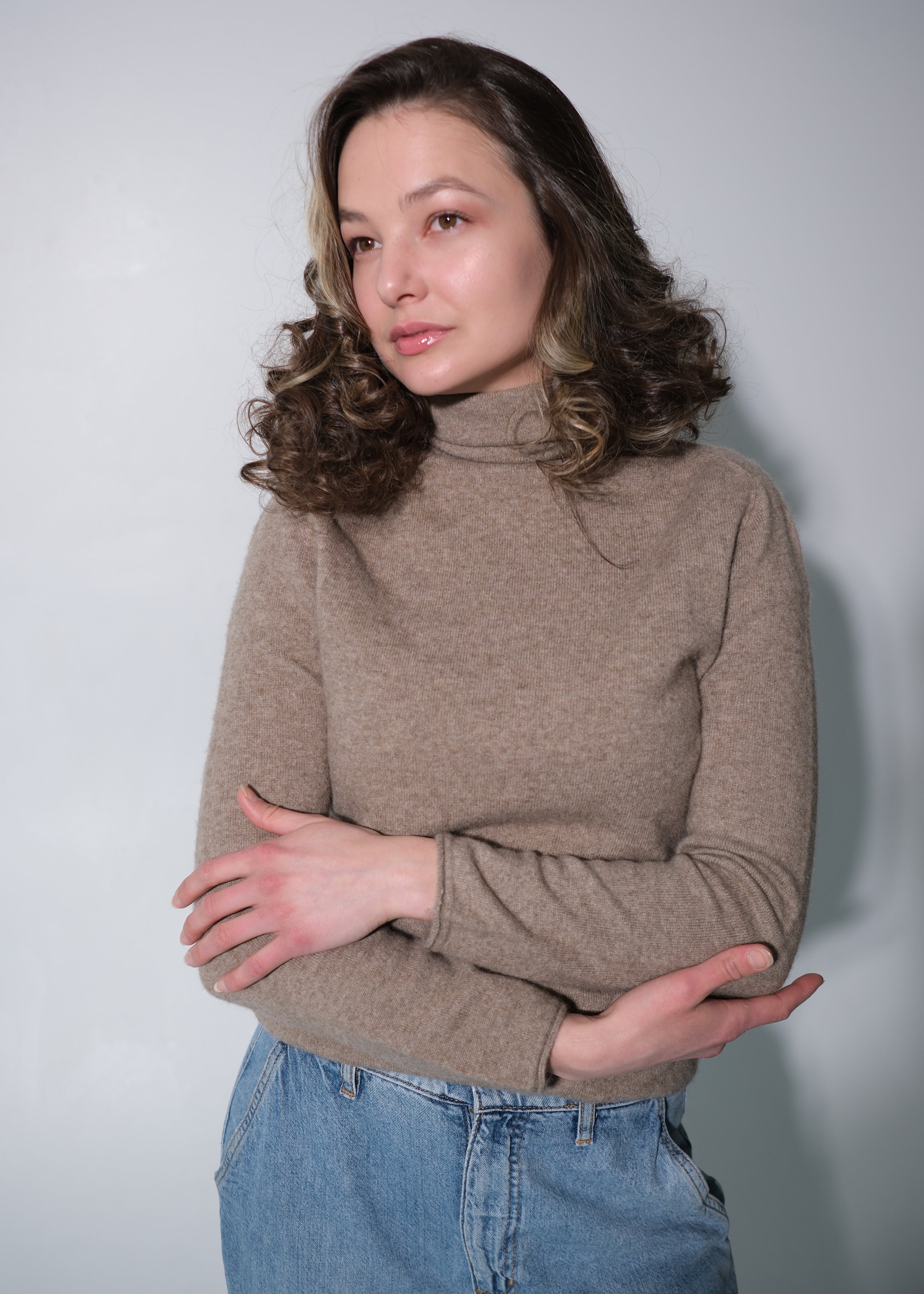 A woman with wavy brown hair wearing a beige turtleneck sweater and blue jeans, standing against a plain light gray background.