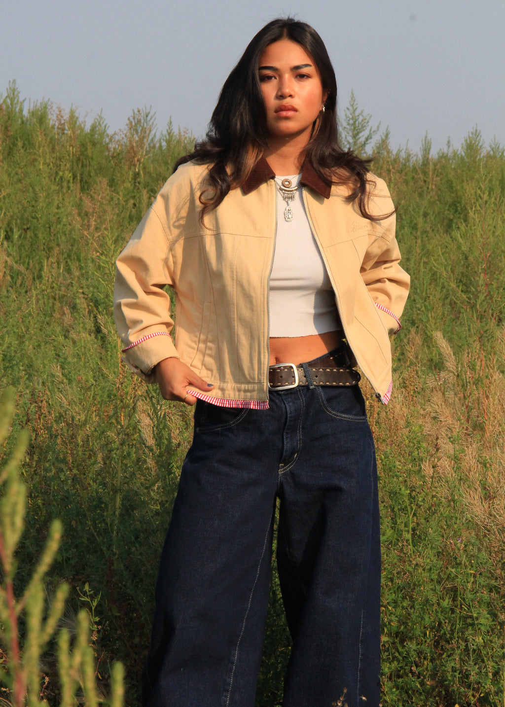 A woman stands outdoors in a grassy field wearing a light beige cropped jacket with a brown collar, a white crop top, dark blue wide-leg jeans, and a black belt.