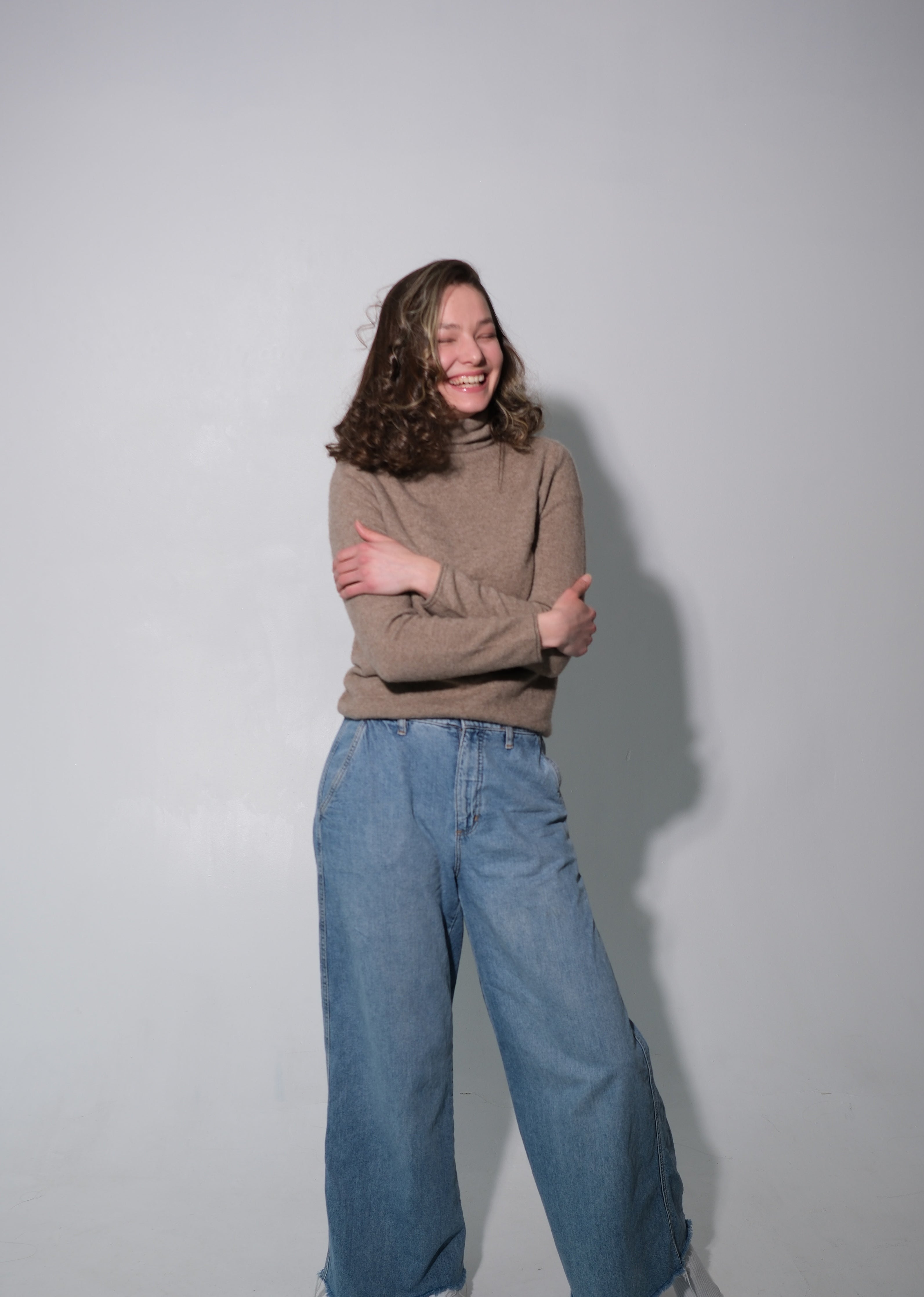 A woman posing in a studio setting, wearing a brown ribbed turtleneck sweater and wide-leg blue denim jeans with white socks.