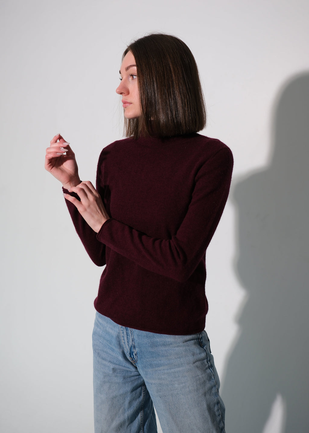 A woman with shoulder-length dark brown hair is shown from the side, wearing a dark burgundy mock-neck long-sleeve knit sweater and light-wash blue jeans, standing against a plain light gray wall with a soft shadow.