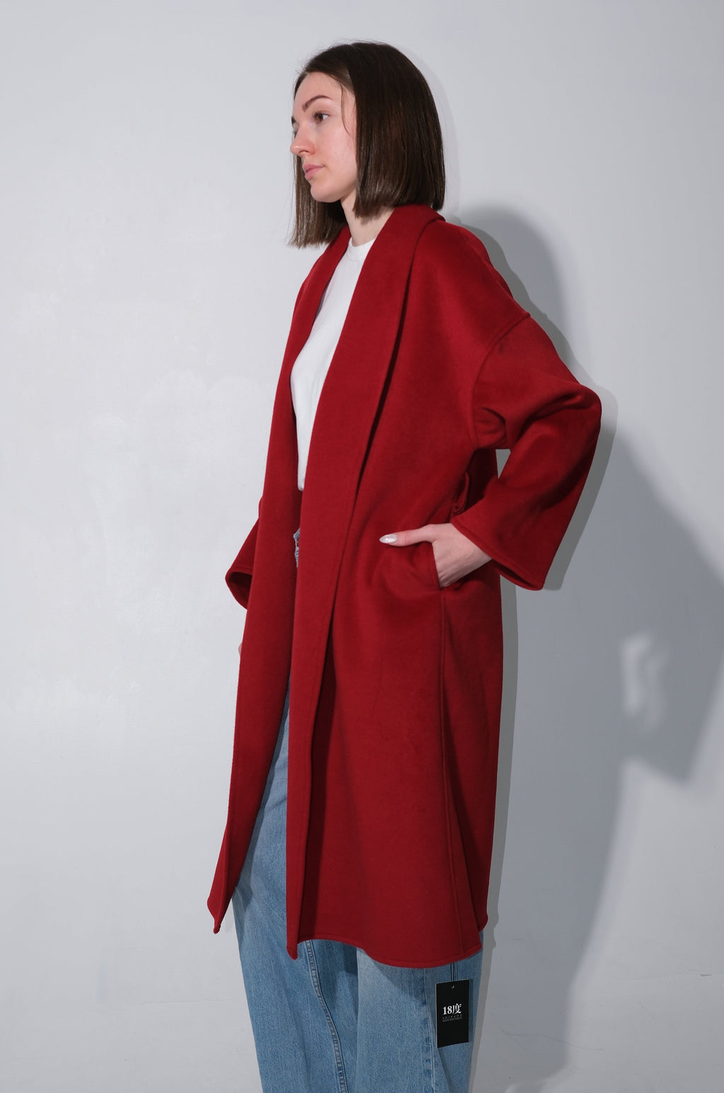 A woman modeling a long, open-front red coat over a white top and light blue jeans, standing against a plain light gray studio background.