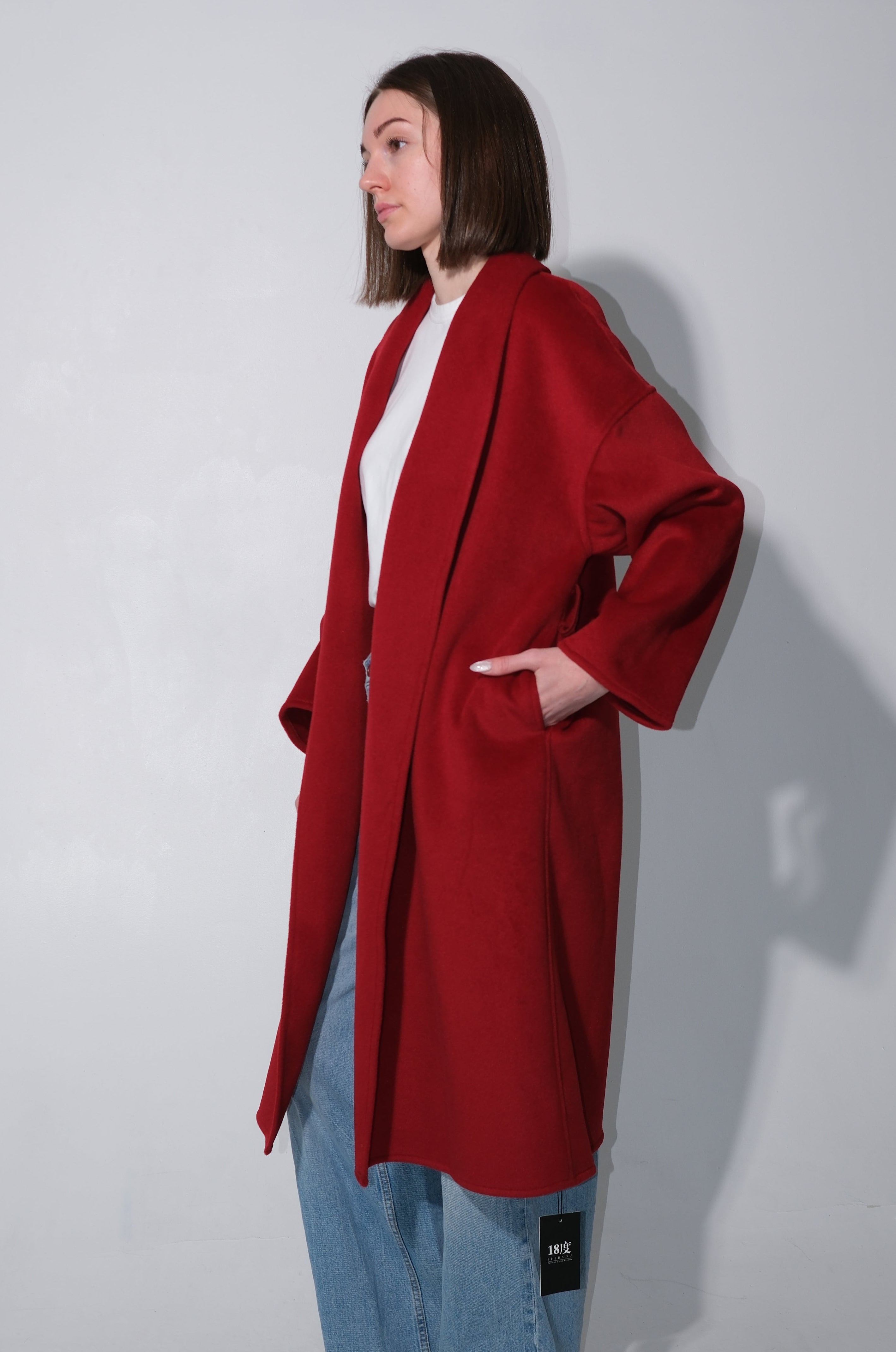 A woman modeling a long, open-front red coat over a white top and light blue jeans, standing against a plain light gray studio background.