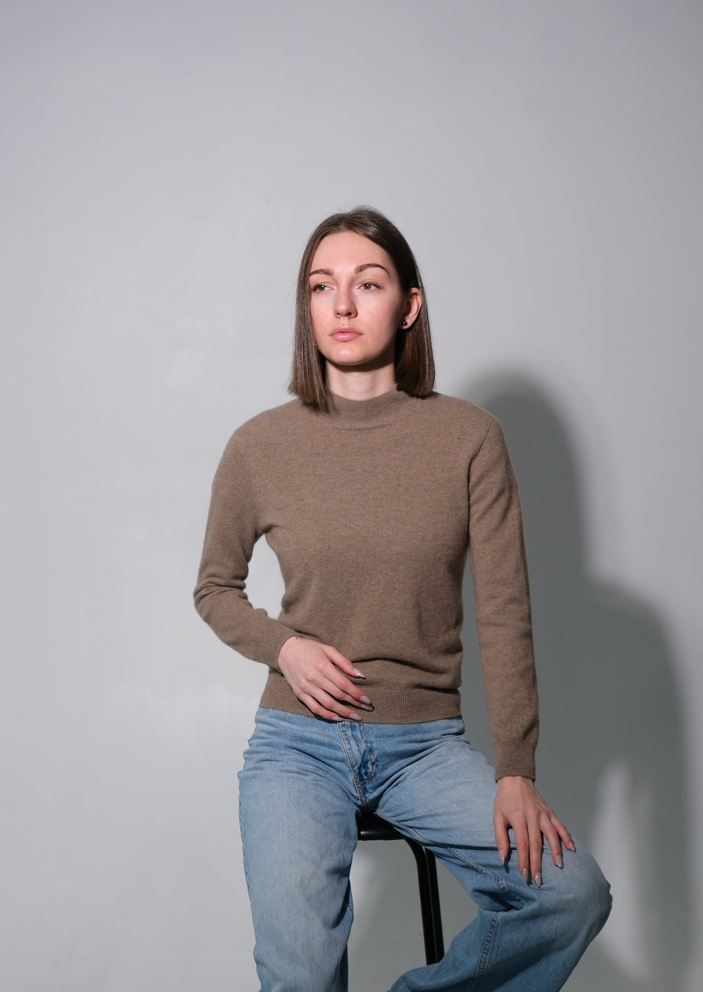 A woman wearing a brown mock-neck knit sweater and light blue jeans sits on a black stool against a gray studio background.