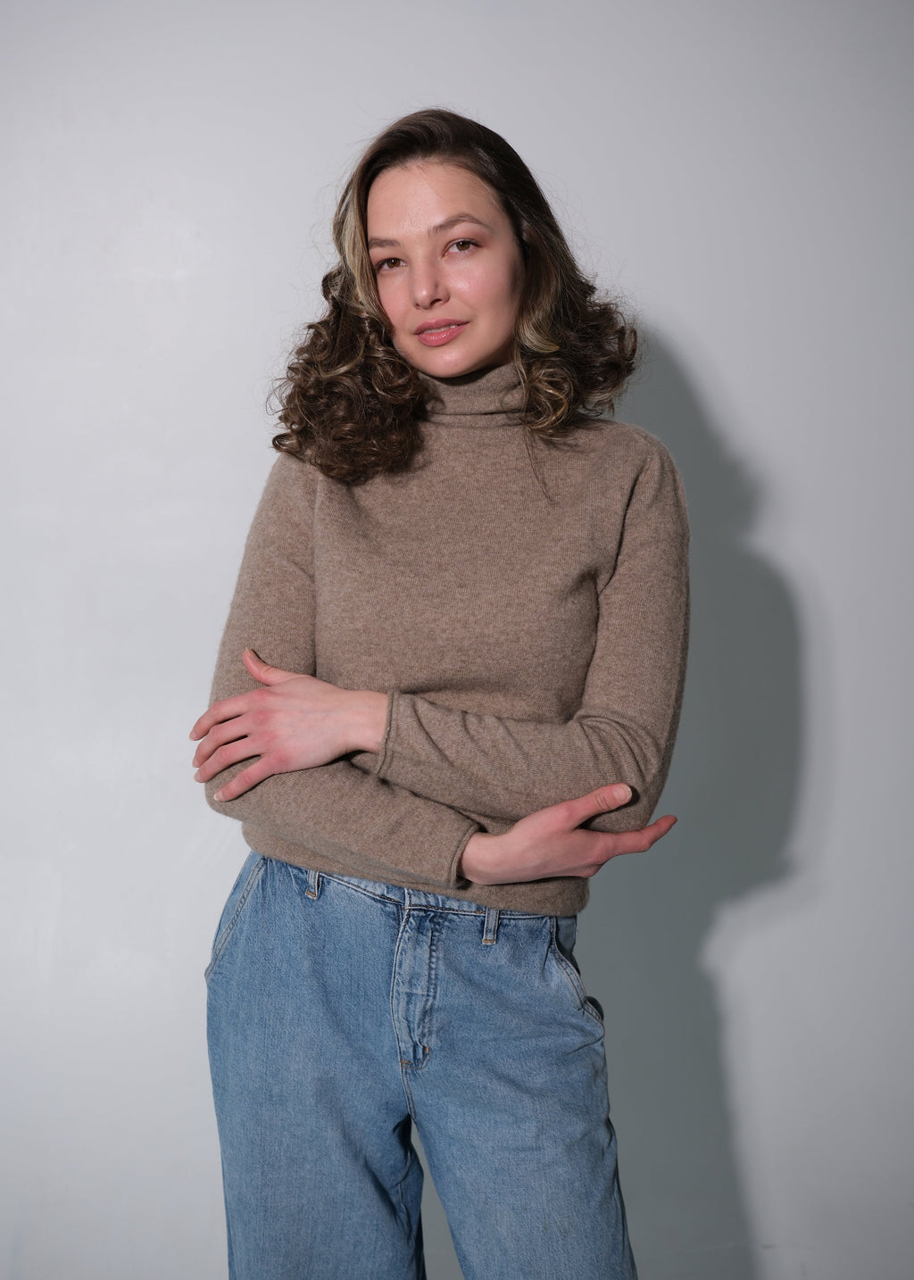A woman with wavy brown hair wearing a beige turtleneck sweater and light blue jeans, standing against a plain light gray wall with her arms crossed.