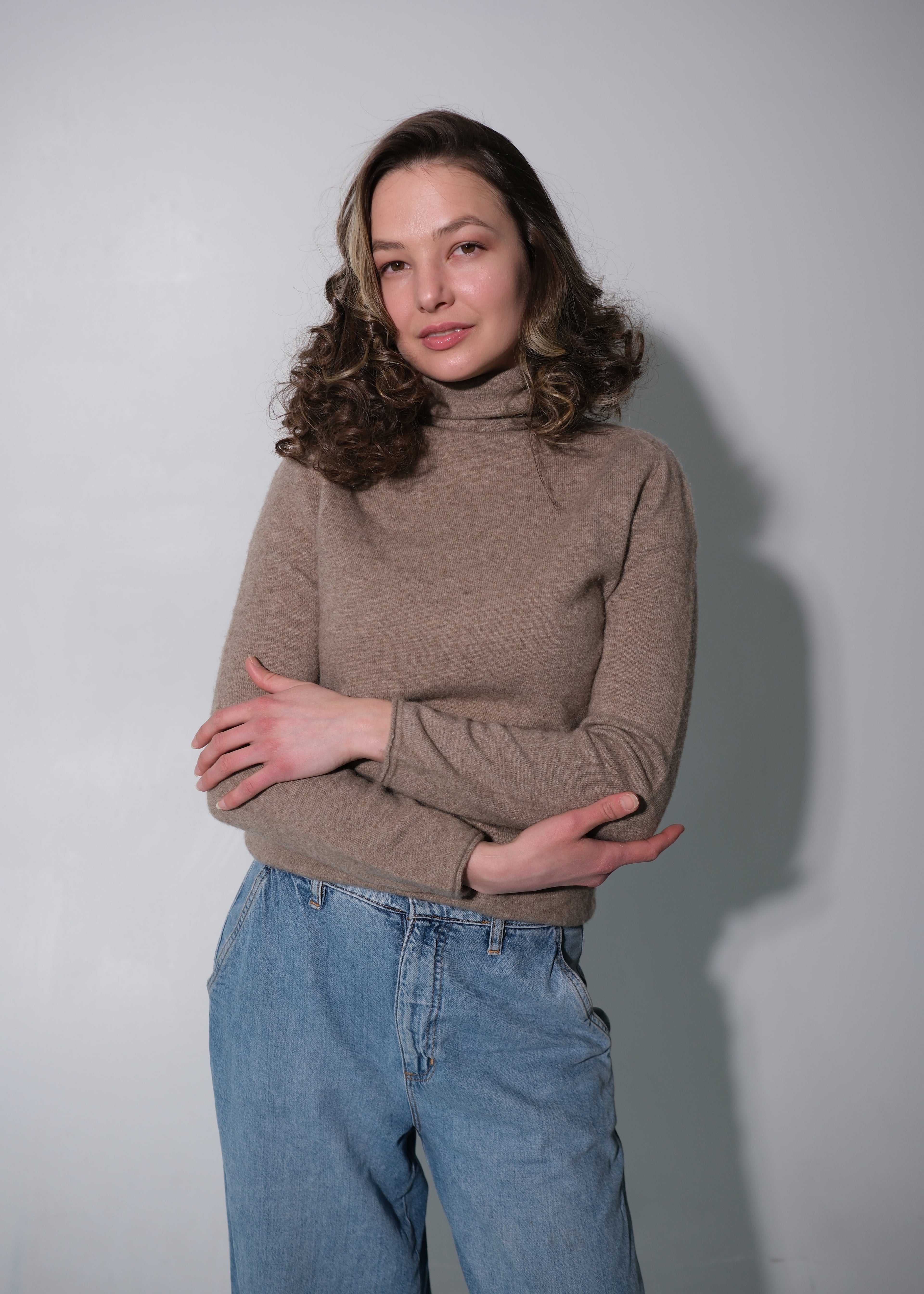 A woman with wavy brown hair wearing a beige turtleneck sweater and light blue jeans, standing against a plain light gray wall with her arms crossed.