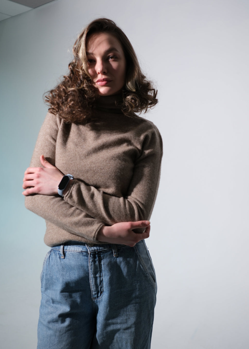 A woman with curly brown hair wearing a beige turtleneck sweater and blue jeans, standing with her arms crossed against a plain light gray wall.