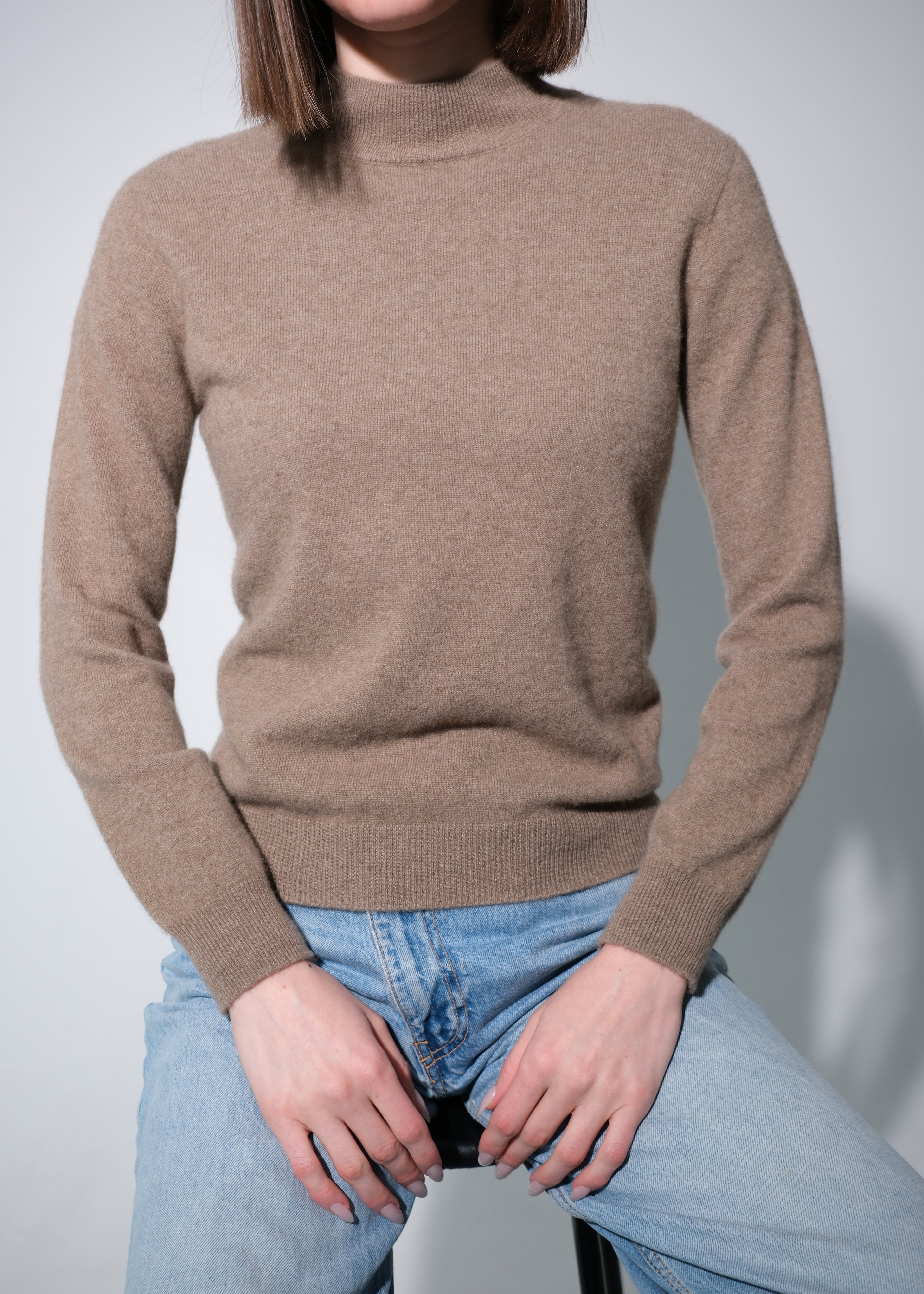 A woman wearing a light brown ribbed mock neck sweater and light blue jeans, sitting on a stool against a light gray background.
