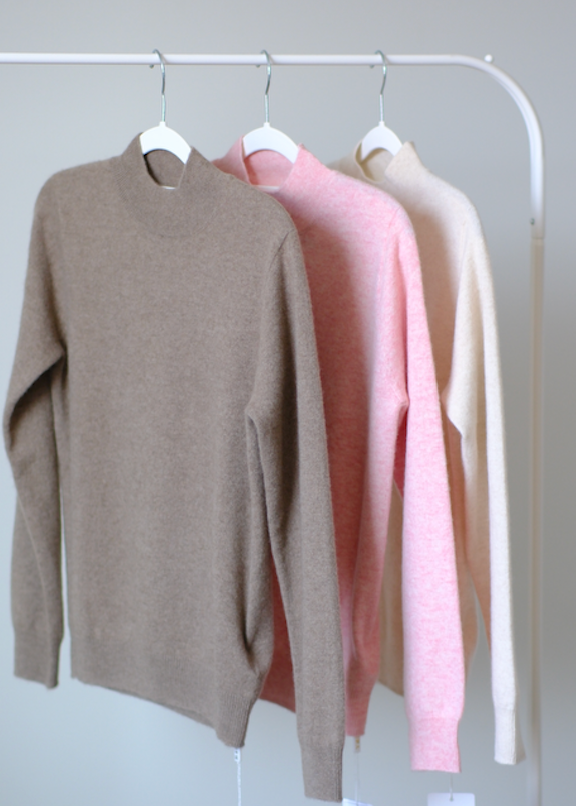 Three knit crewneck sweaters in taupe, light pink, and cream hang on white hangers from a white clothing rack against a plain light gray wall.