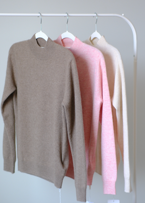 Three knit crewneck sweaters in taupe, light pink, and cream hang on white hangers from a white clothing rack against a plain light gray wall.