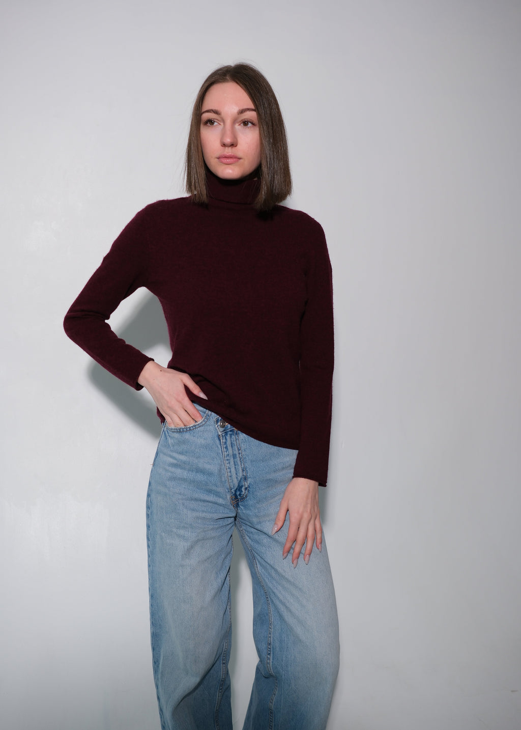 A woman modeling a dark burgundy ribbed turtleneck sweater paired with light-wash blue jeans, standing against a plain light gray studio background.