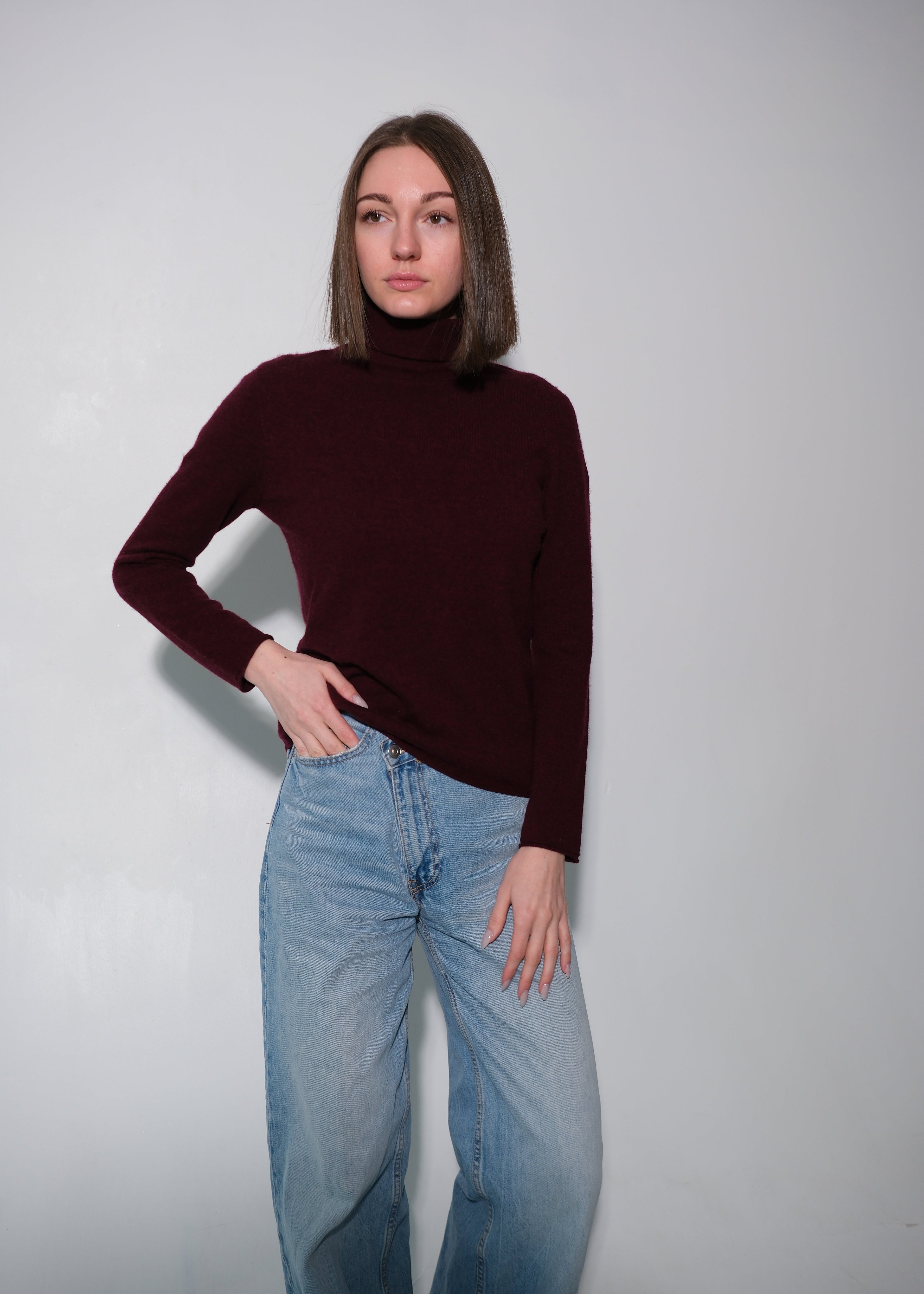 A woman modeling a dark burgundy ribbed turtleneck sweater paired with light-wash blue jeans, standing against a plain light gray studio background.