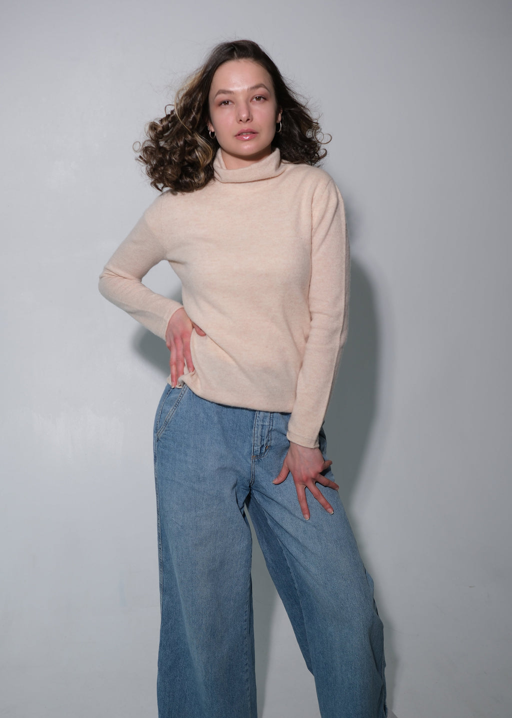 A woman posing in a light beige mock-neck knit sweater and wide-leg blue jeans against a plain light gray studio background.