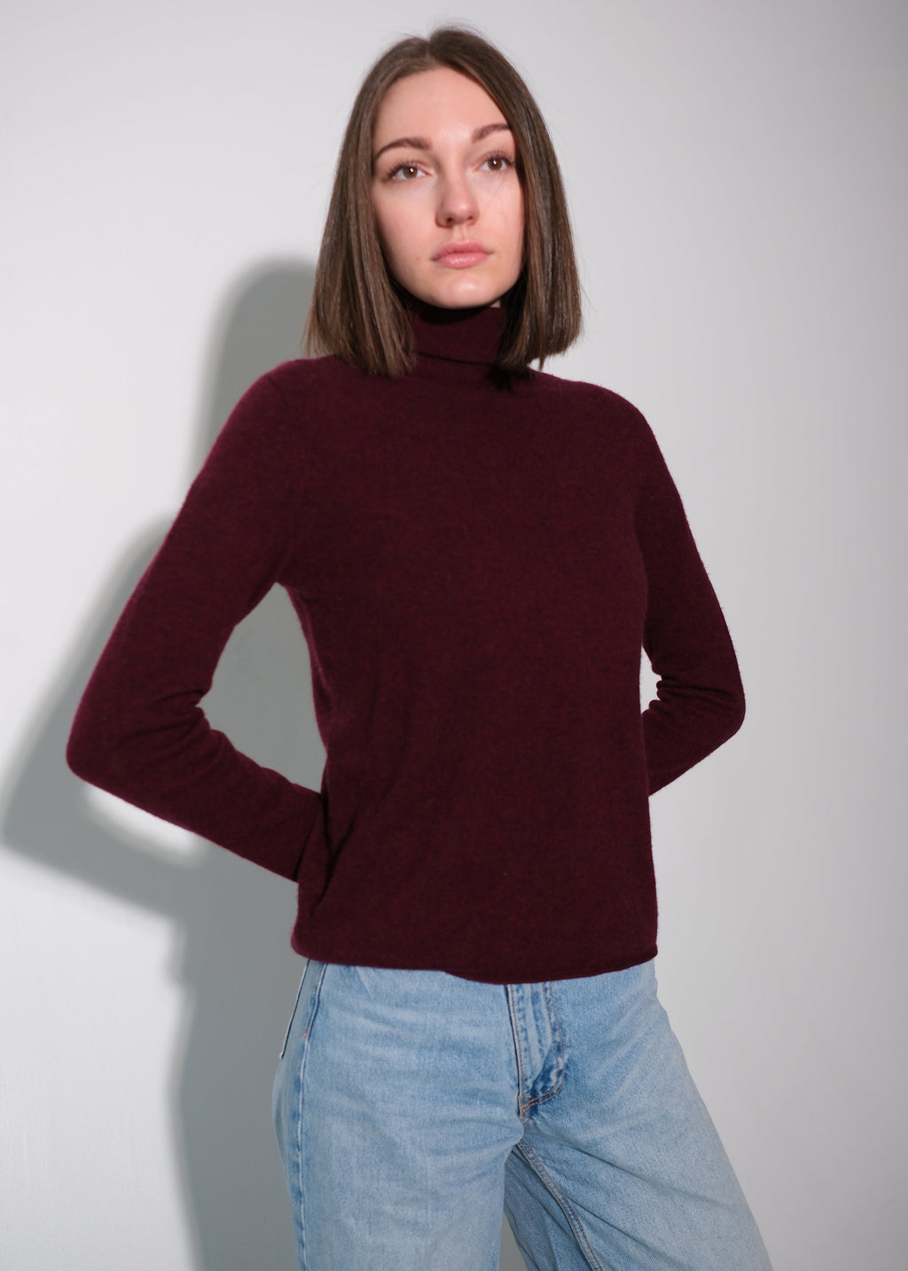A woman with shoulder-length brown hair wearing a dark burgundy mock neck knit sweater and light blue jeans, standing against a light gray wall.