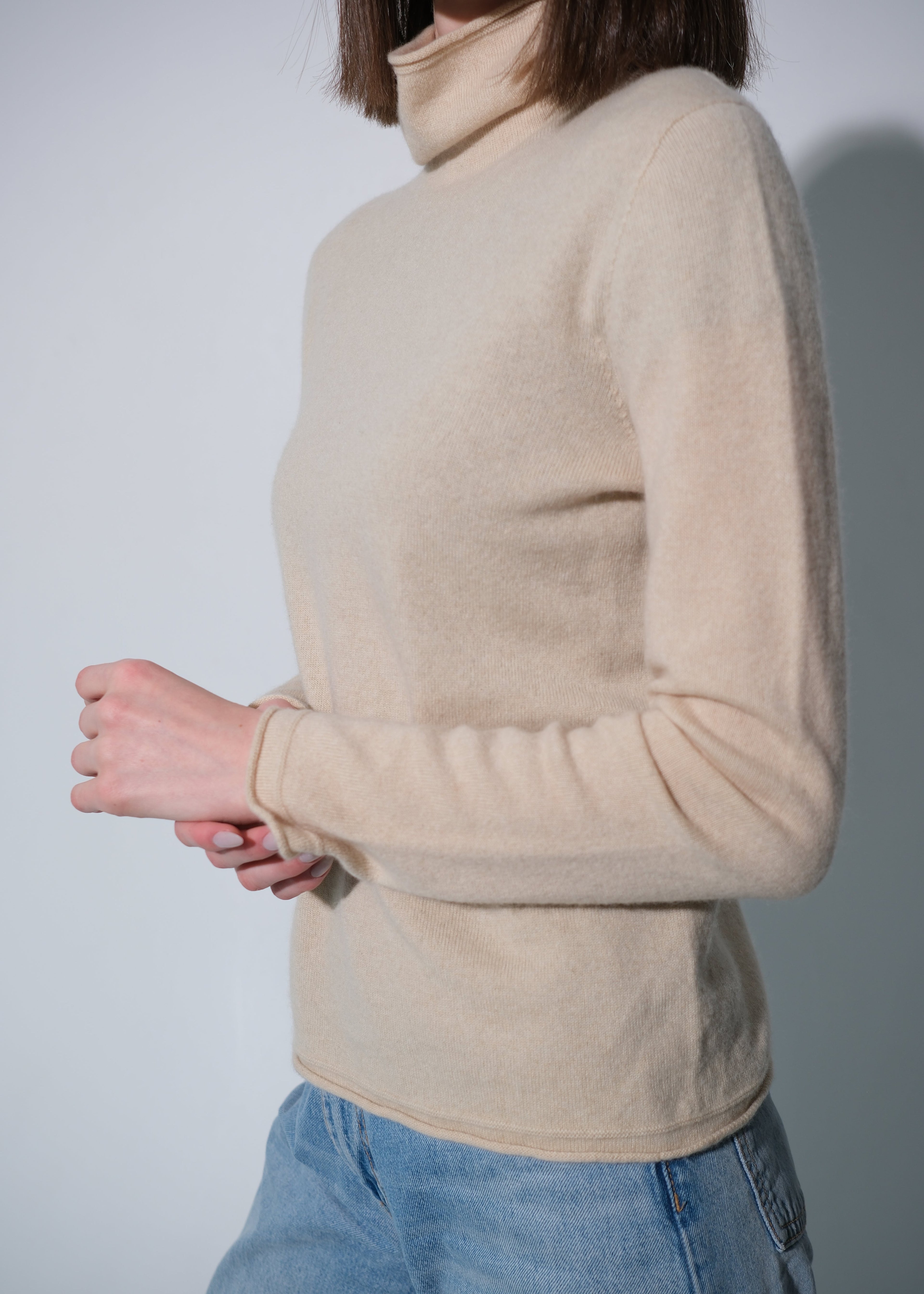 A person wearing a light beige turtleneck sweater and blue jeans, shown from the side against a plain light gray background.