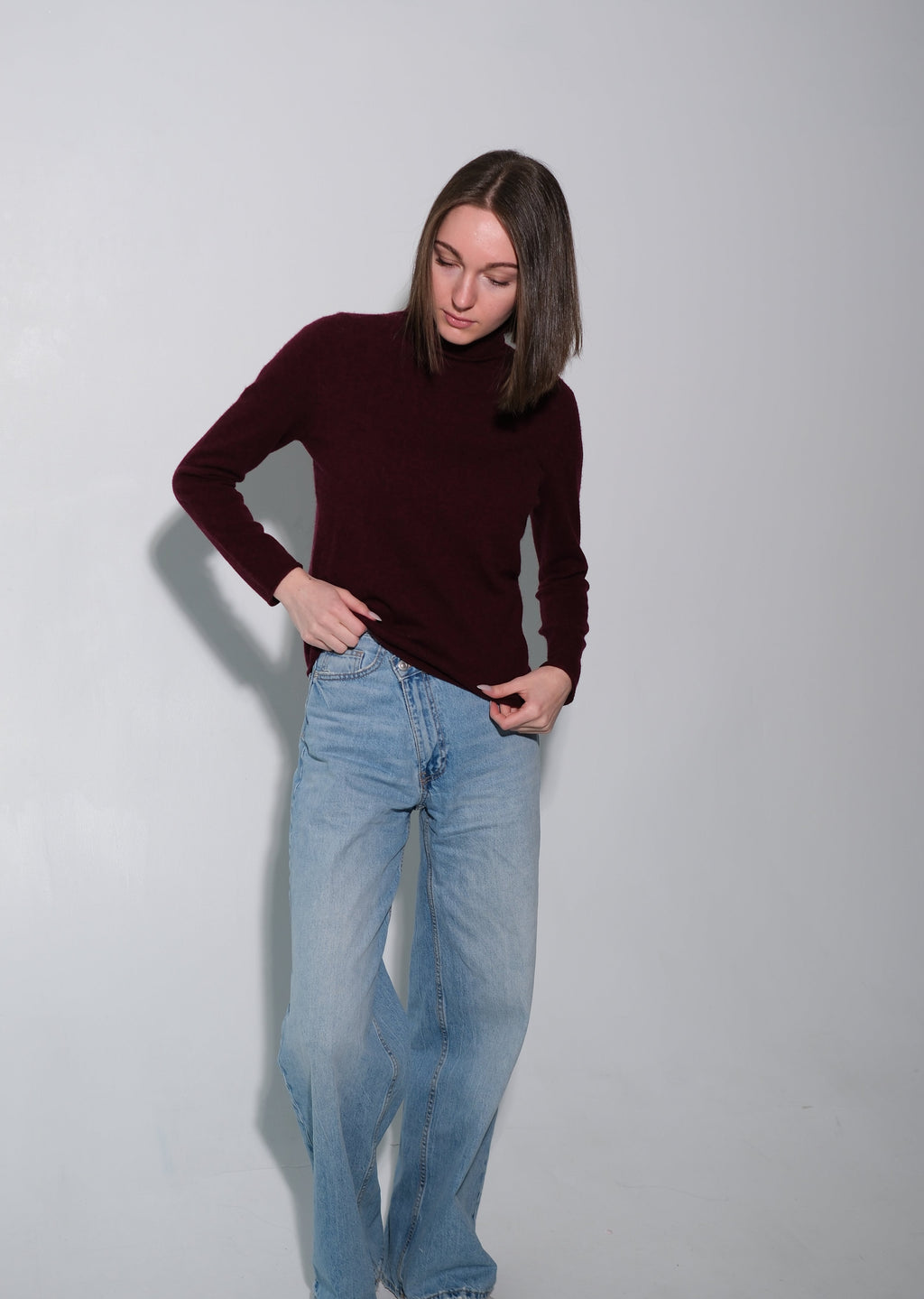 A woman modeling a dark burgundy long-sleeve mock neck sweater and light blue wide-leg jeans against a plain light gray studio background.
