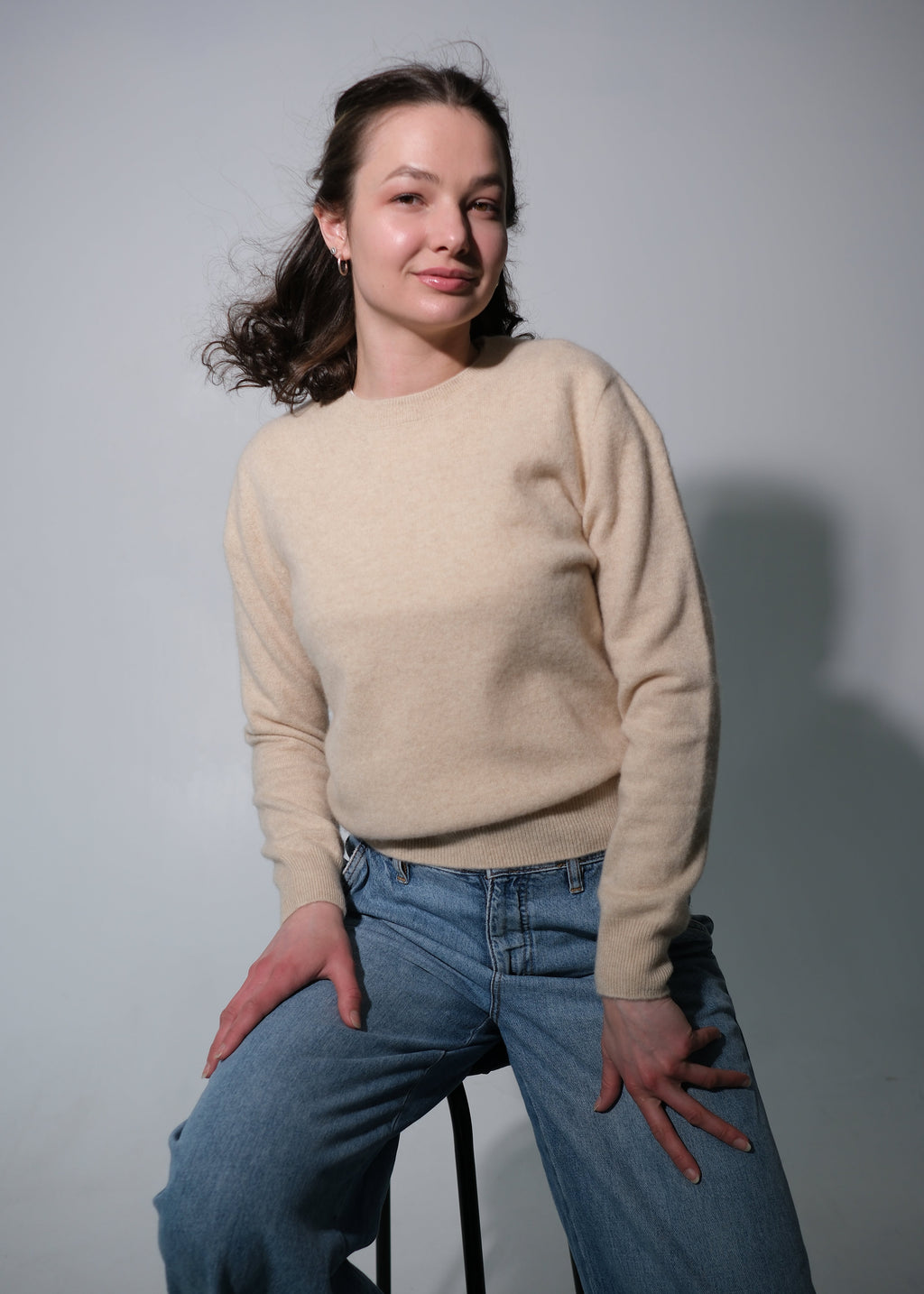 A woman wearing a beige crewneck knit sweater and blue jeans, sitting on a black stool against a light gray studio background.