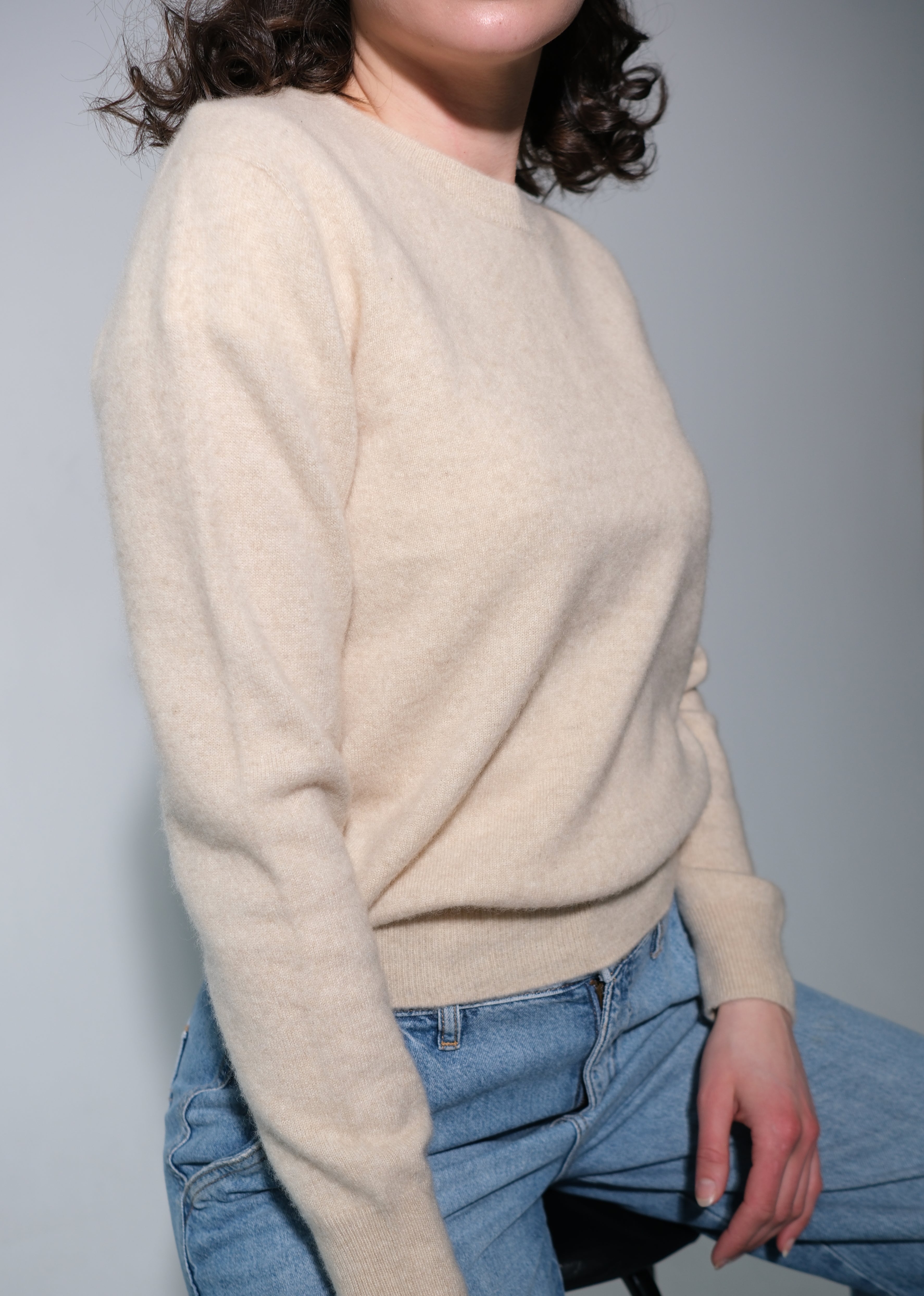 A person wearing a cream-colored knit sweater and light blue jeans, seated on a black stool against a plain light gray wall.